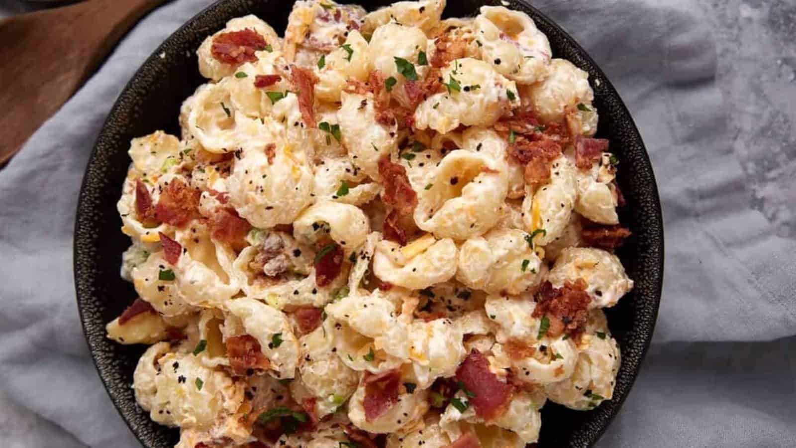Shell pasta salad with creamy dressing, bacon bits, and chopped herbs in a black bowl on a gray cloth.