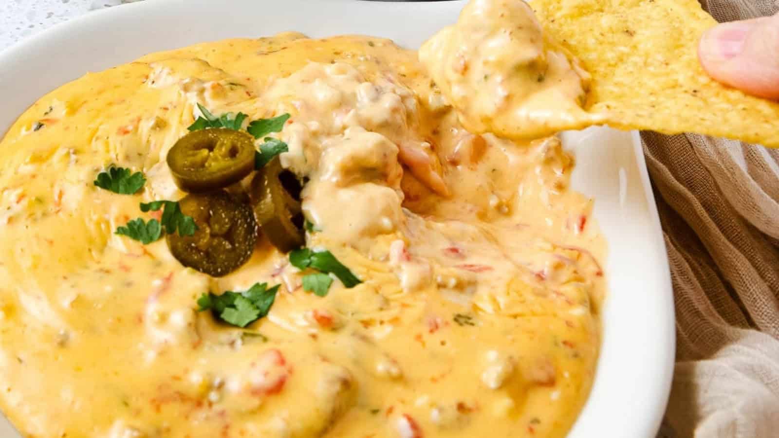 Creamy queso dip topped with jalapeños and parsley, with a tortilla chip being dipped into the bowl.