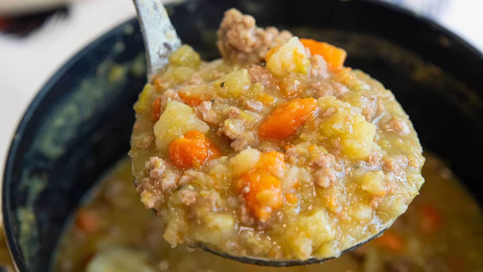 A close-up of a ladle holding chunky stew with ground meat, carrots, and potatoes over a pot.