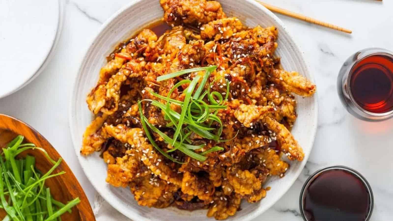 A plate of crispy fried chicken topped with sesame seeds and sliced green onions, with drinks on the side.
