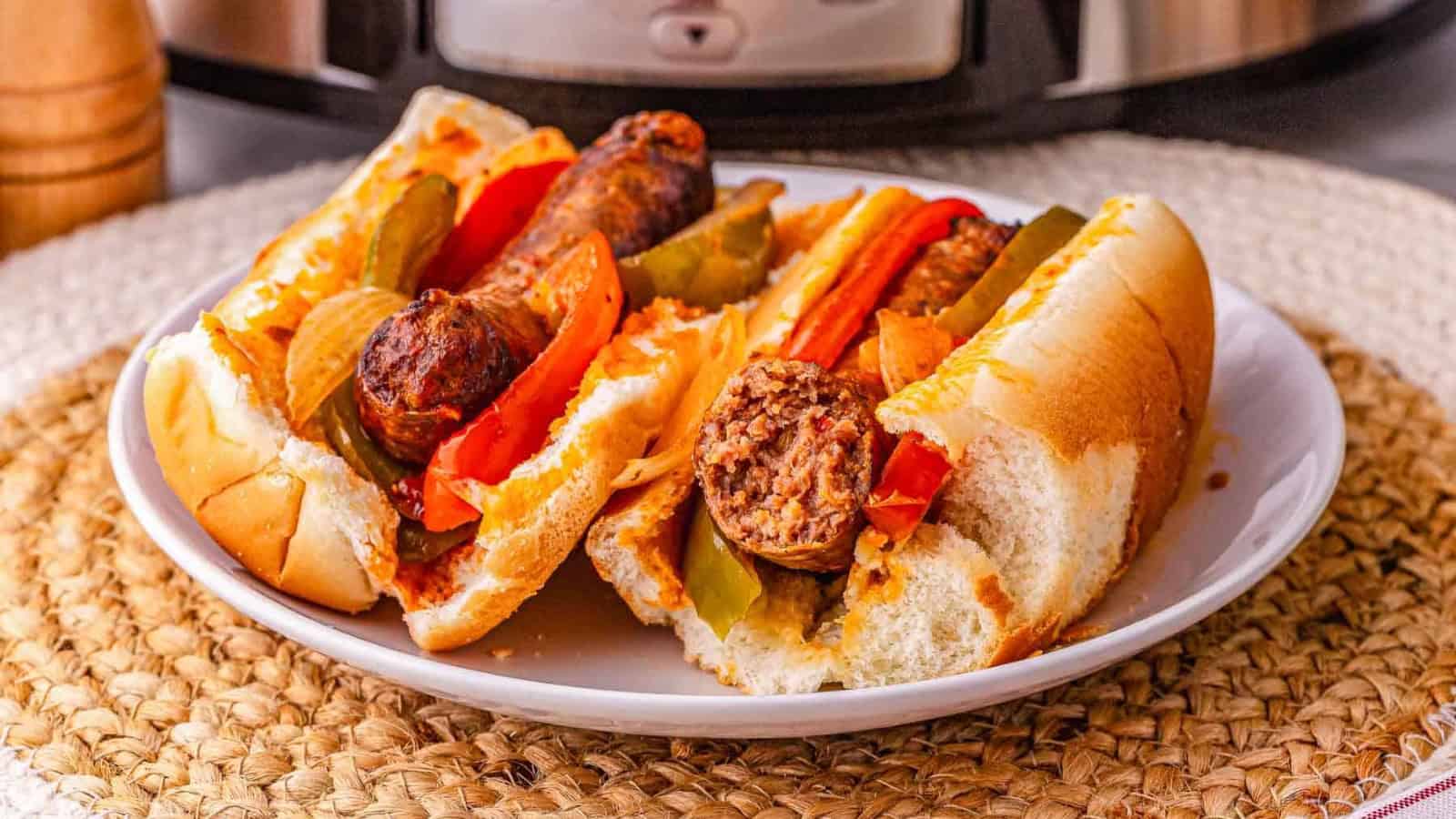 Two sausage sandwiches with peppers and onions on a white plate, sitting on a woven mat.