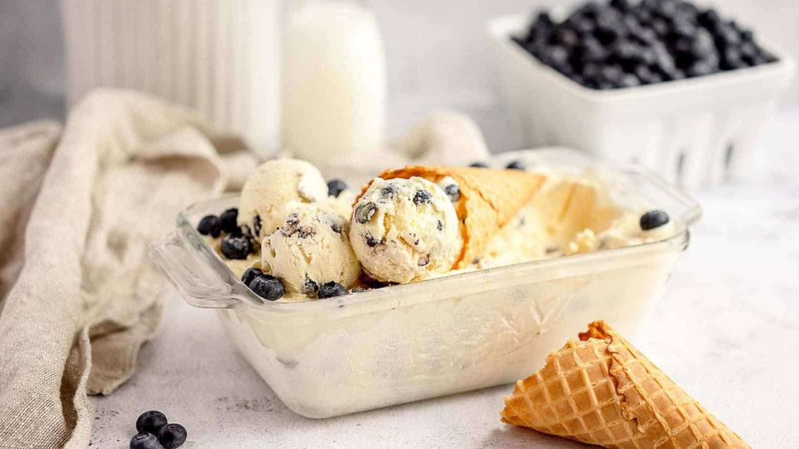 Vanilla ice cream with blueberries and a waffle cone in a glass dish, with a milk jug and blueberries in the background.