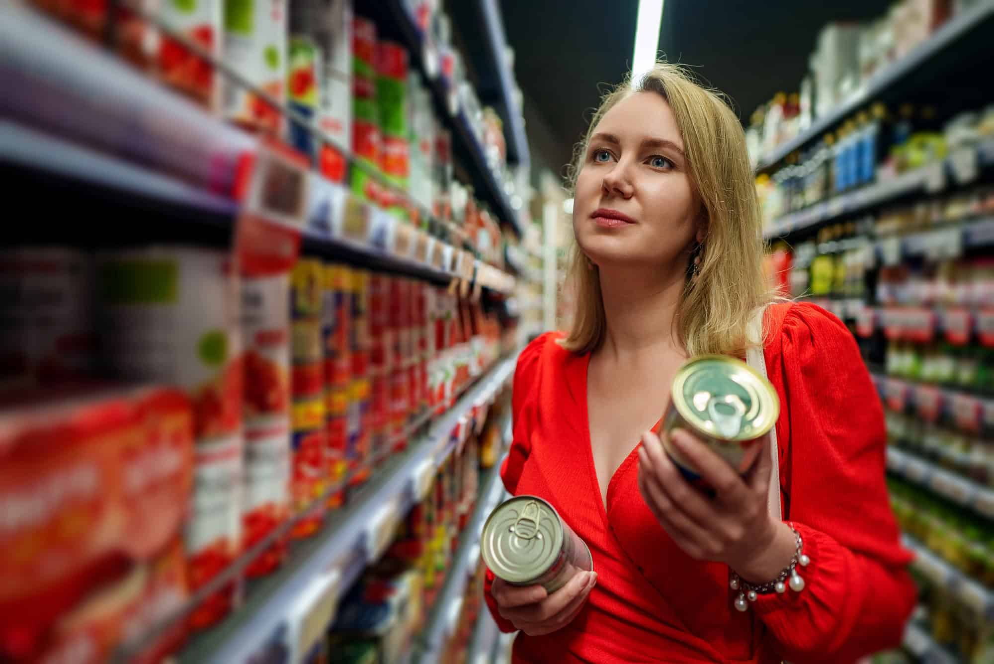 Woman in a red dress holding canned goods and shopping in a grocery store aisle.