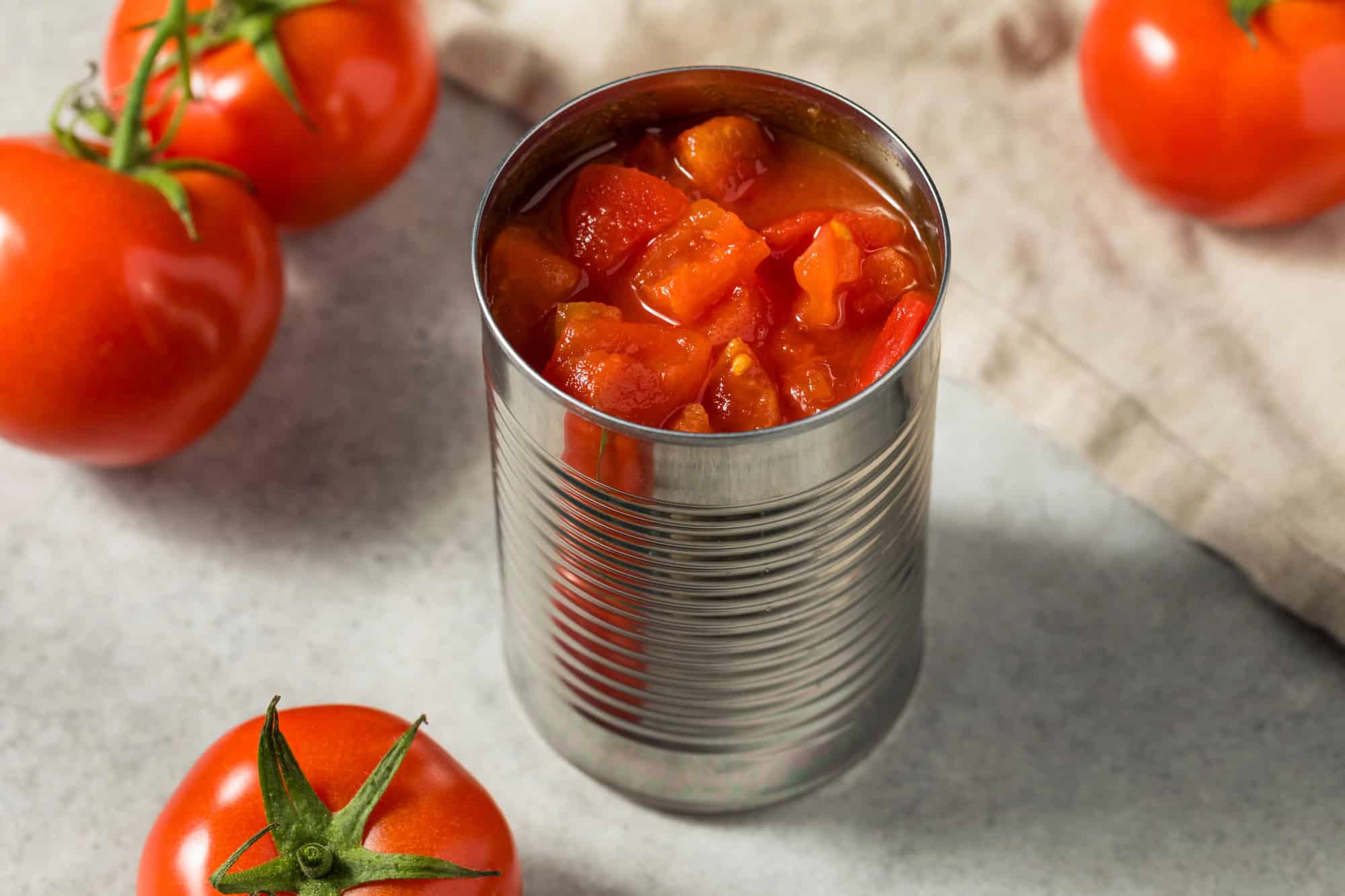 An open can of diced tomatoes surrounded by fresh whole tomatoes on a light surface.