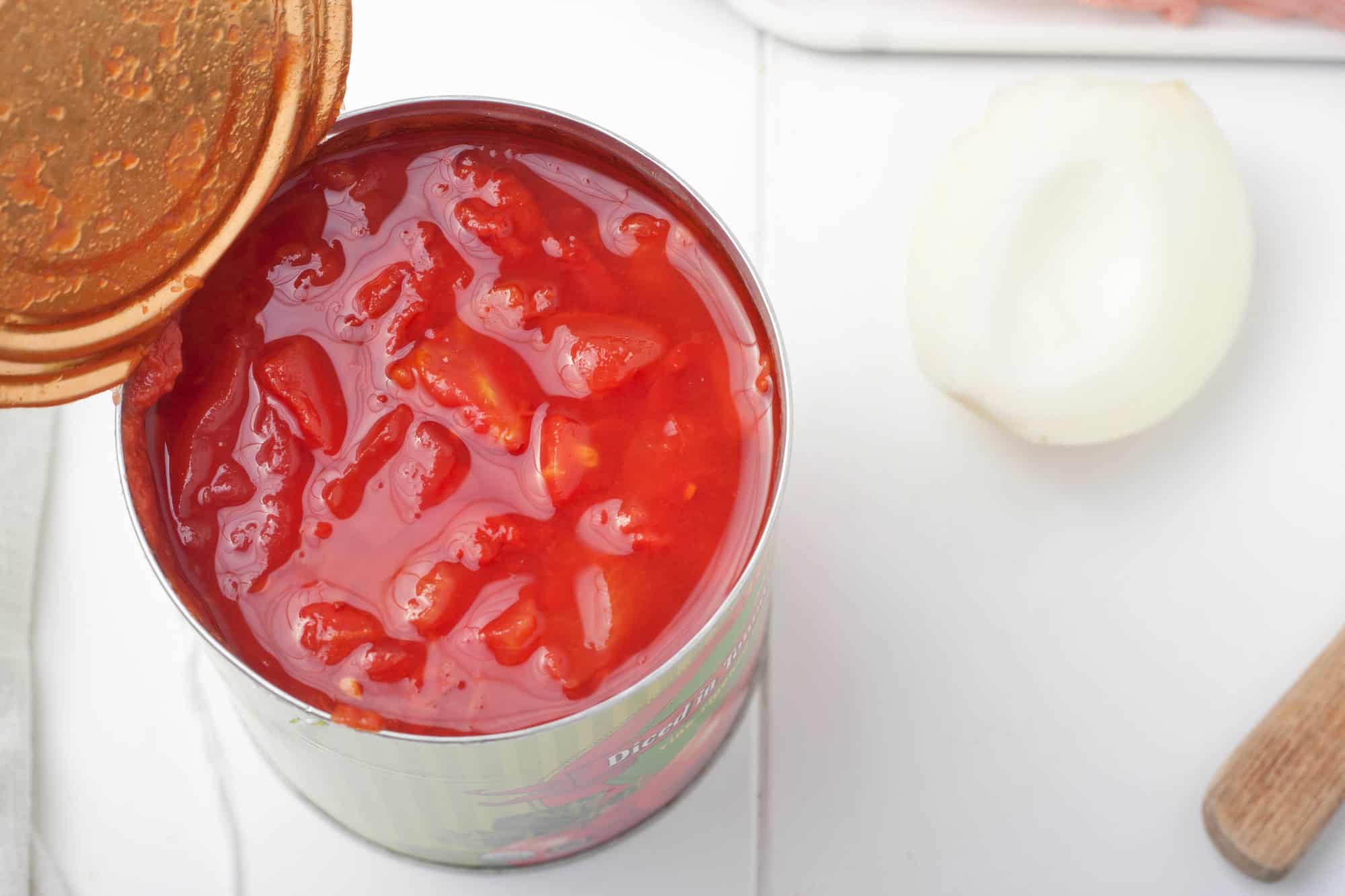 Open can of diced tomatoes next to a halved onion and a wooden utensil on a white surface.