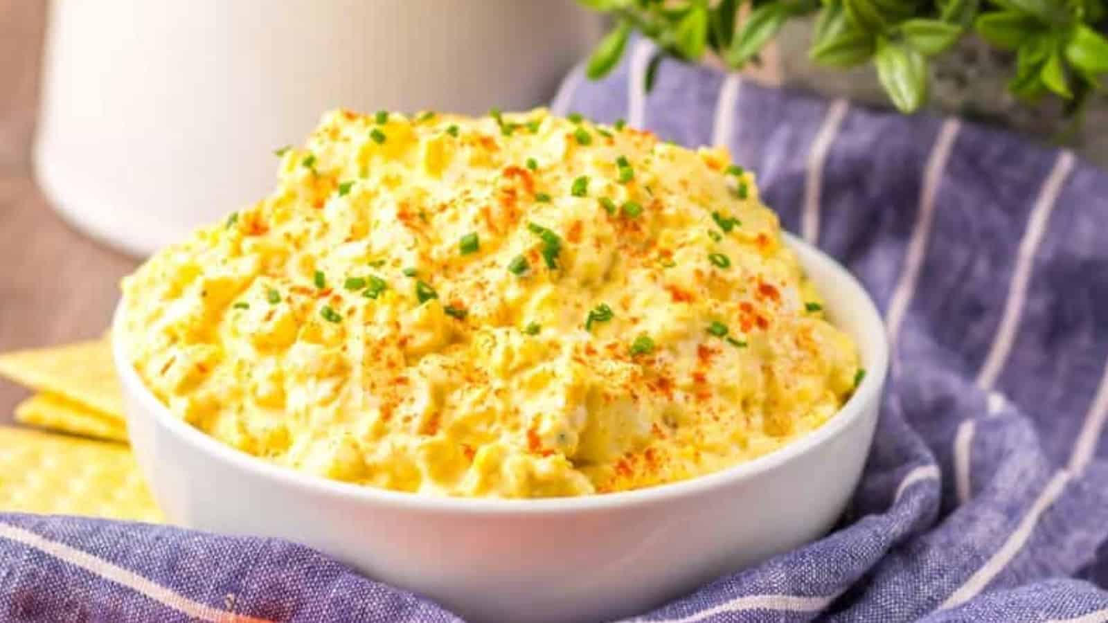 A bowl of deviled egg dip garnished with chives and paprika, surrounded by baby carrots on a striped cloth.