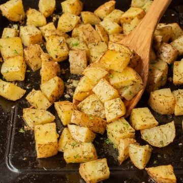 Cubed roasted potatoes with herbs on a baking tray, stirred with a wooden spoon.