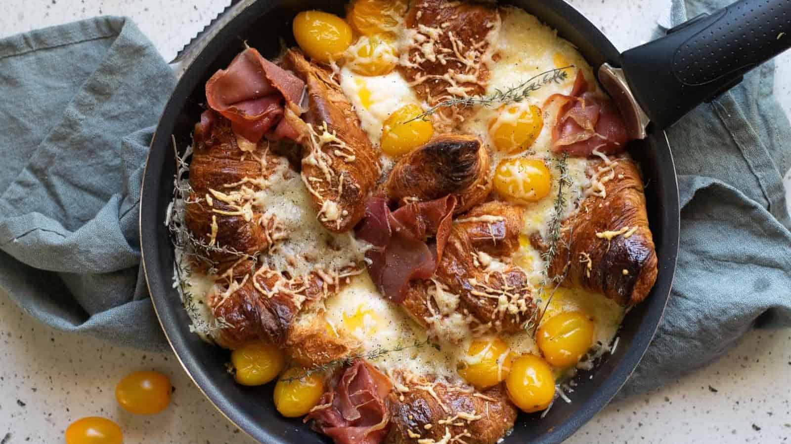 Croissants, yellow tomatoes, ham, cheese, and herbs baked in a skillet on a gray cloth.