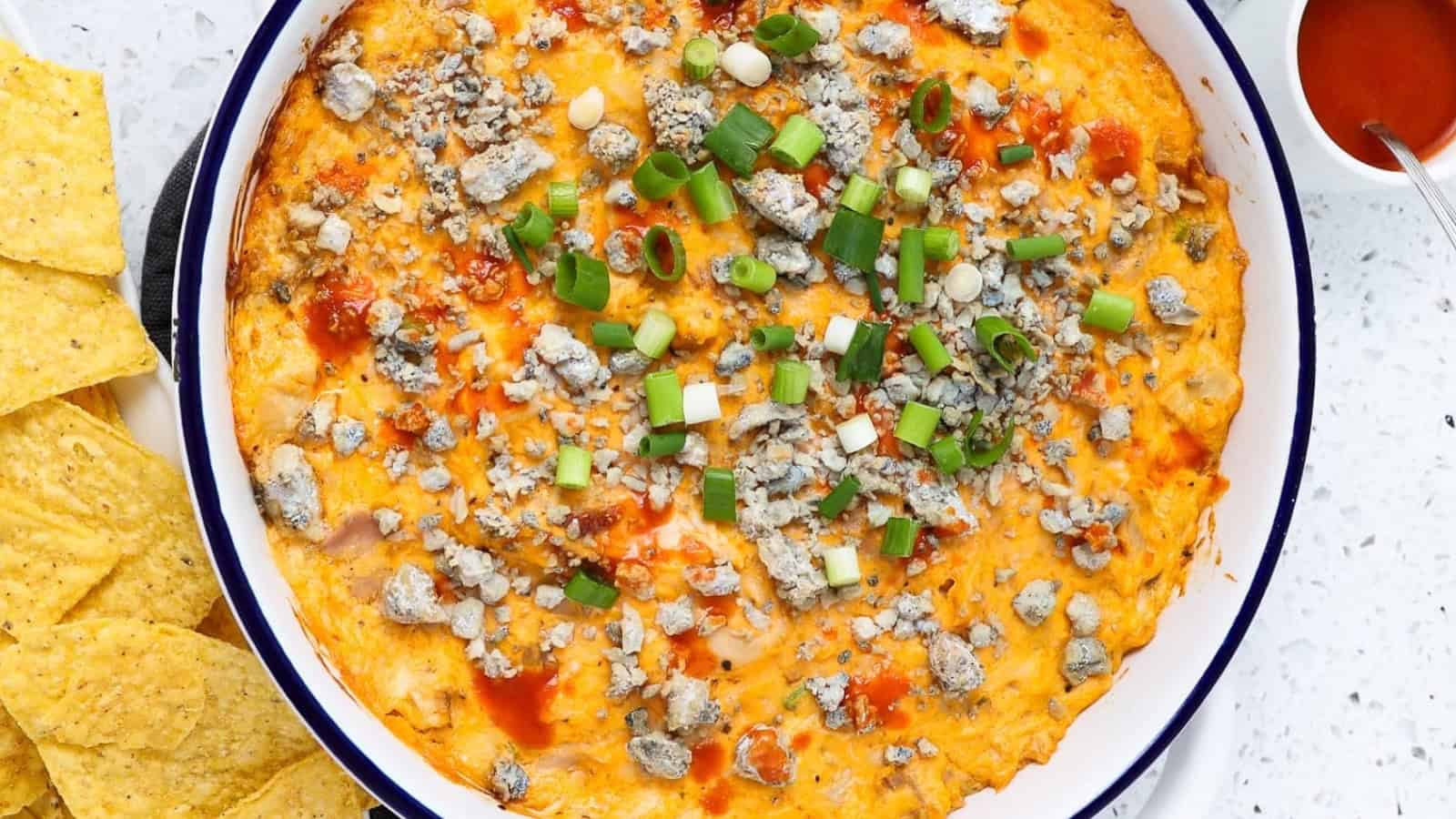 A bowl of buffalo chicken dip topped with blue cheese and green onions, served with tortilla chips.