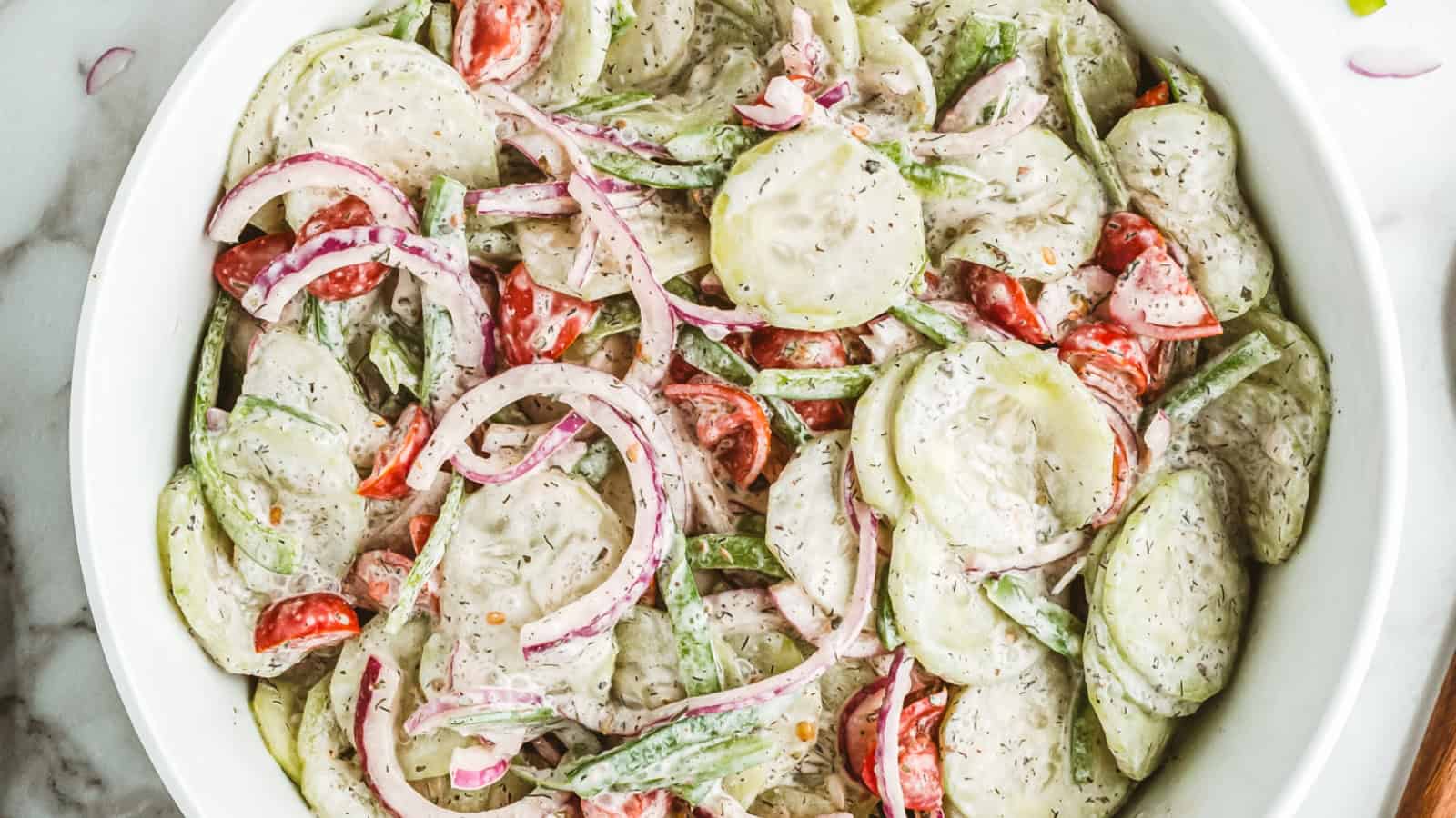 Cucumber salad with red onions, tomatoes, and green peppers in a creamy dressing, served in a white bowl.