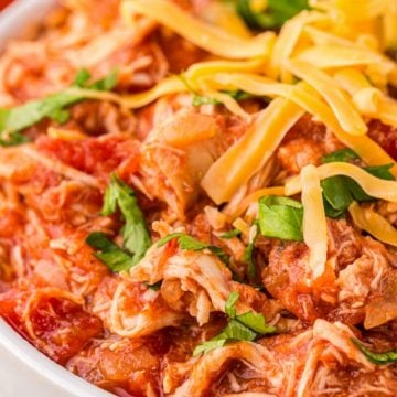 Close-up of shredded chicken in tomato sauce, topped with cheese and fresh herbs in a white bowl.