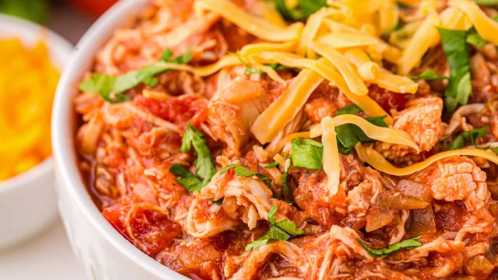 Close-up of shredded chicken in tomato sauce, topped with cheese and fresh herbs in a white bowl.