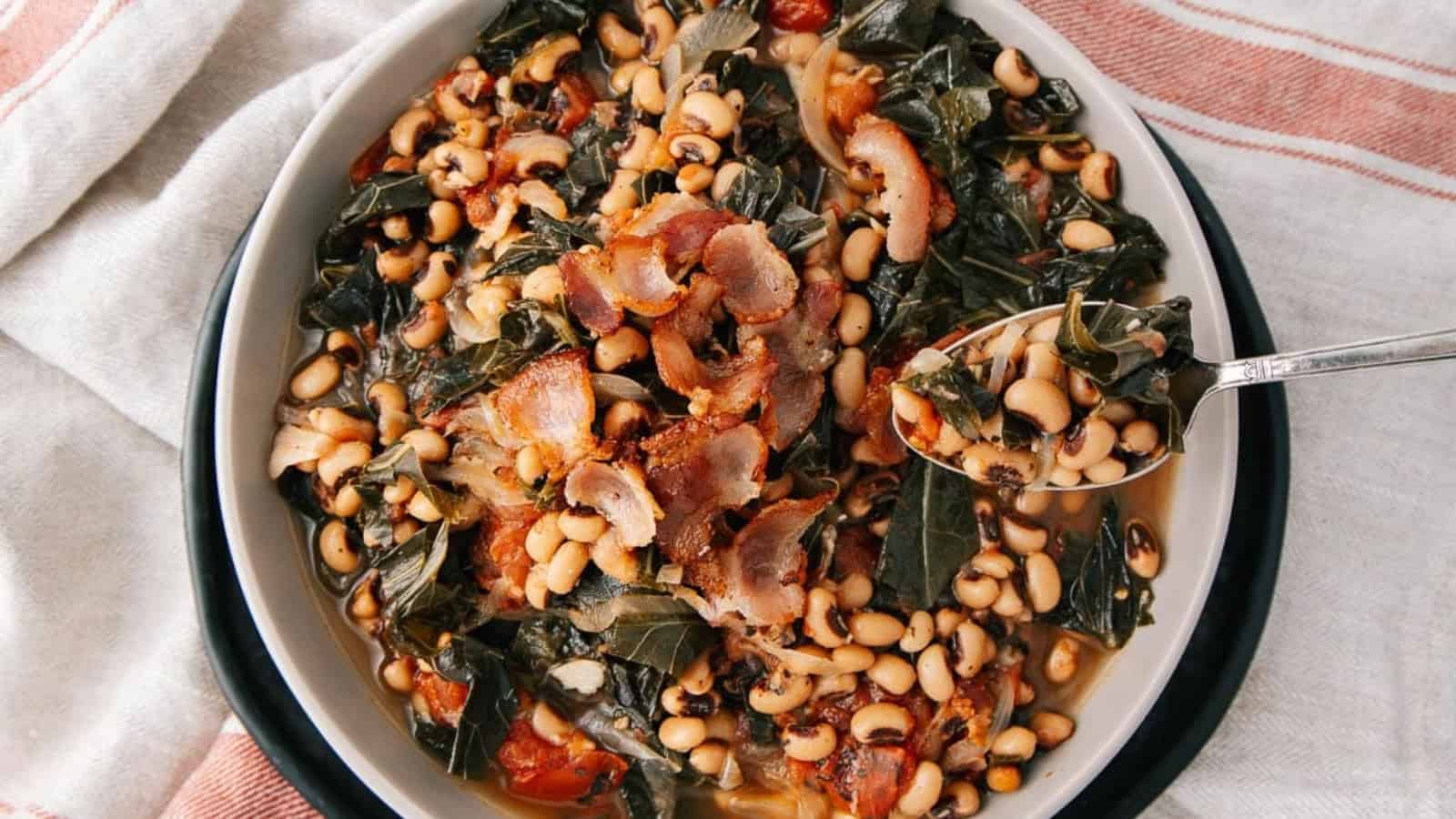 A bowl of black-eyed peas with collard greens, bacon, and tomatoes, with a spoon on the side.