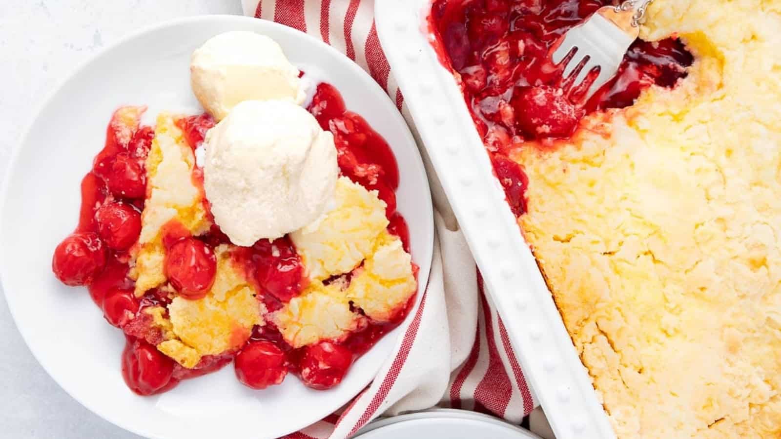 Cherry cobbler with vanilla ice cream on a plate next to a baking dish of cobbler, on a striped towel.