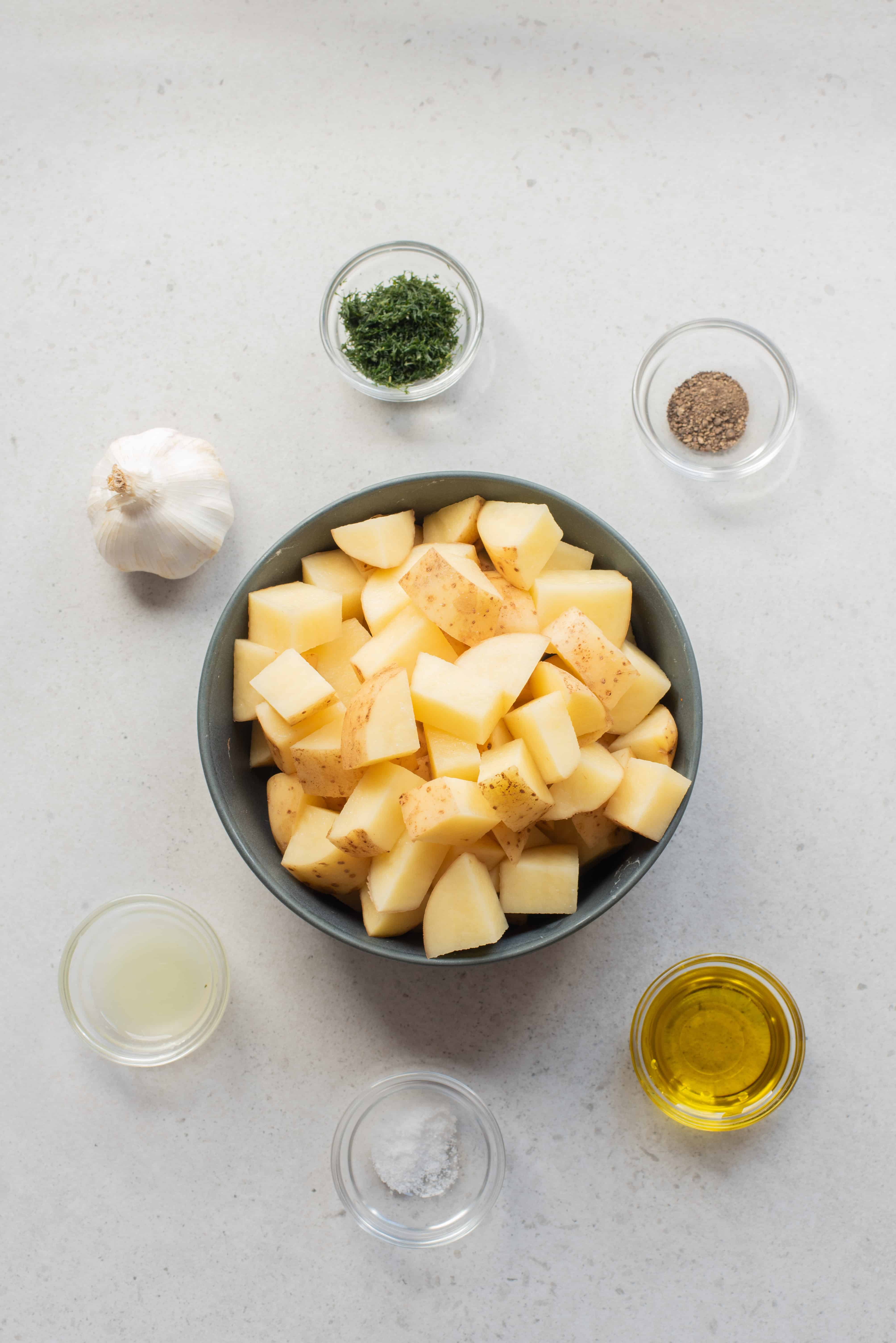 A bowl of chopped potatoes surrounded by garlic, herbs, pepper, oil, lemon juice, and salt on a countertop.