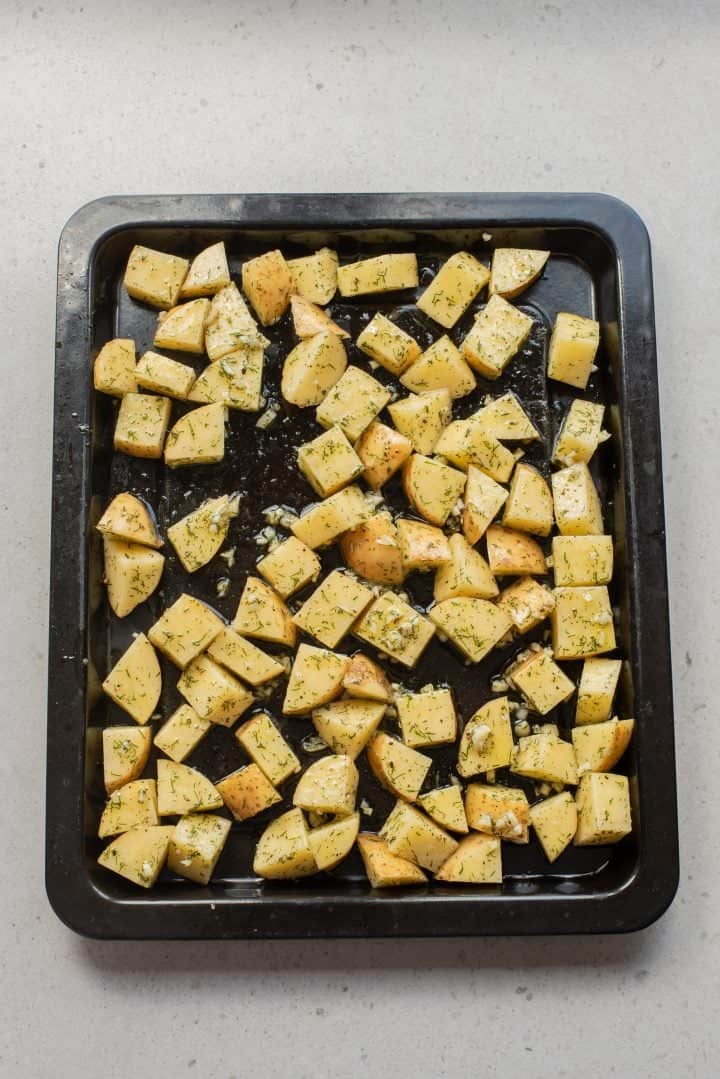 Cubed potatoes seasoned with herbs and oil spread on a black baking tray, ready to be roasted.