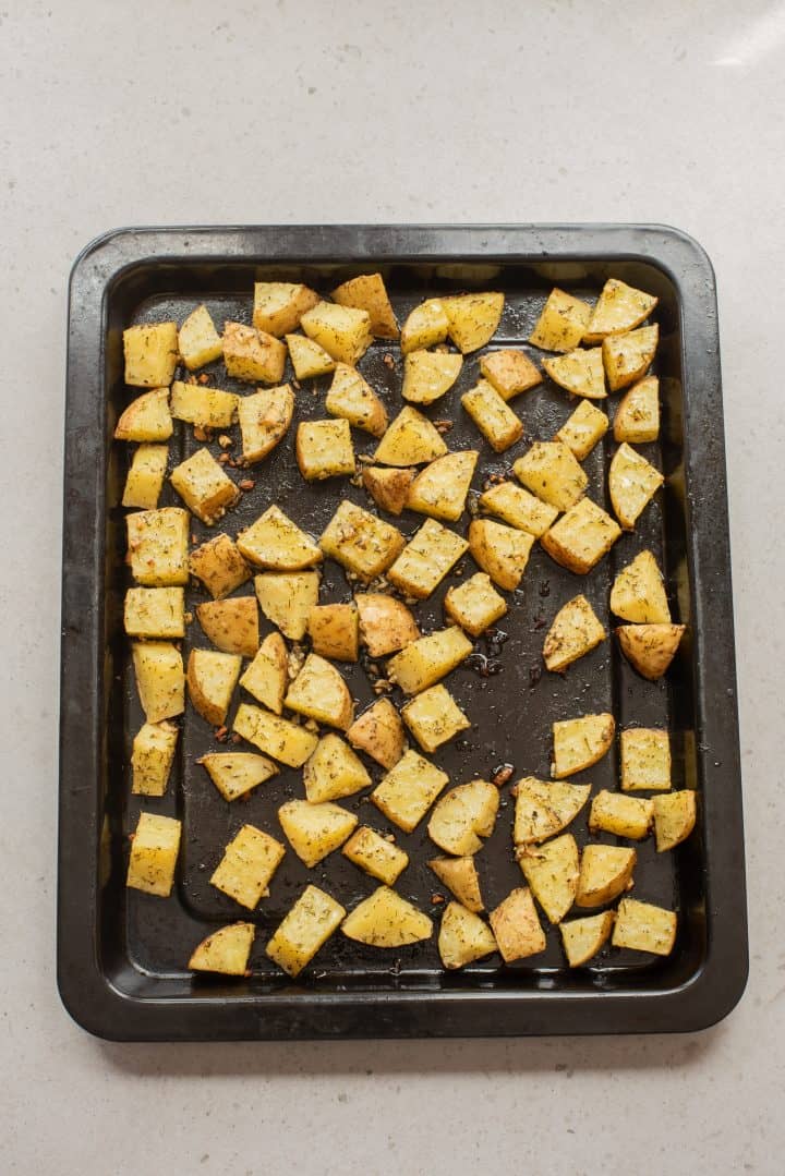 Cubed, seasoned potatoes spread on a black baking tray, ready to be roasted, on a light countertop.