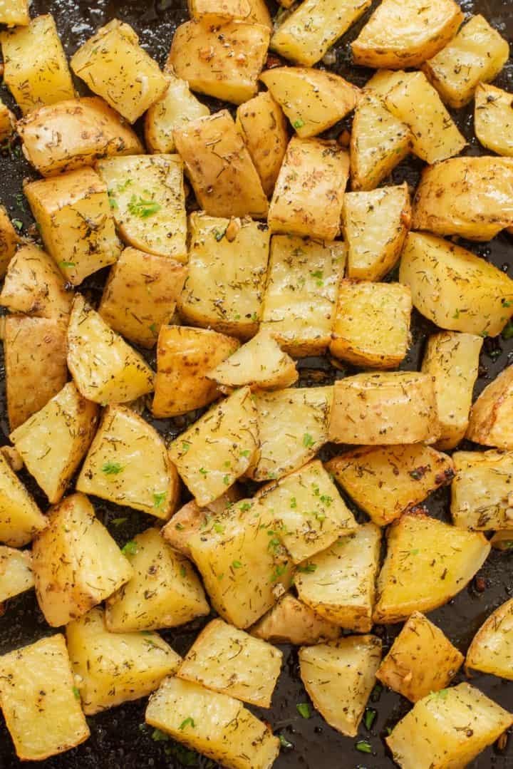 Roasted potato cubes with herbs and seasoning scattered on a dark baking sheet.