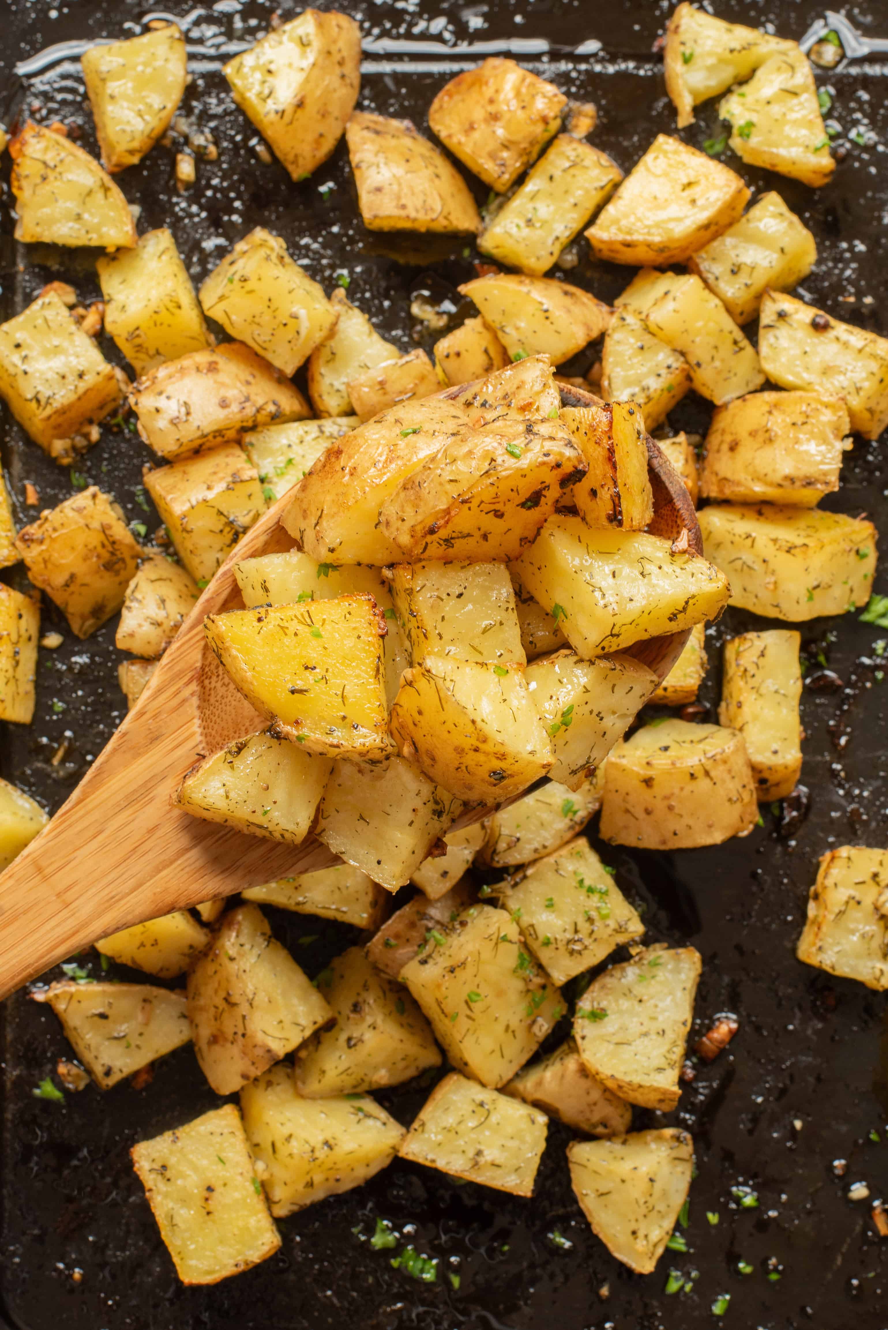 Roasted potato cubes with herbs on a baking sheet, some lifted with a wooden spoon.
