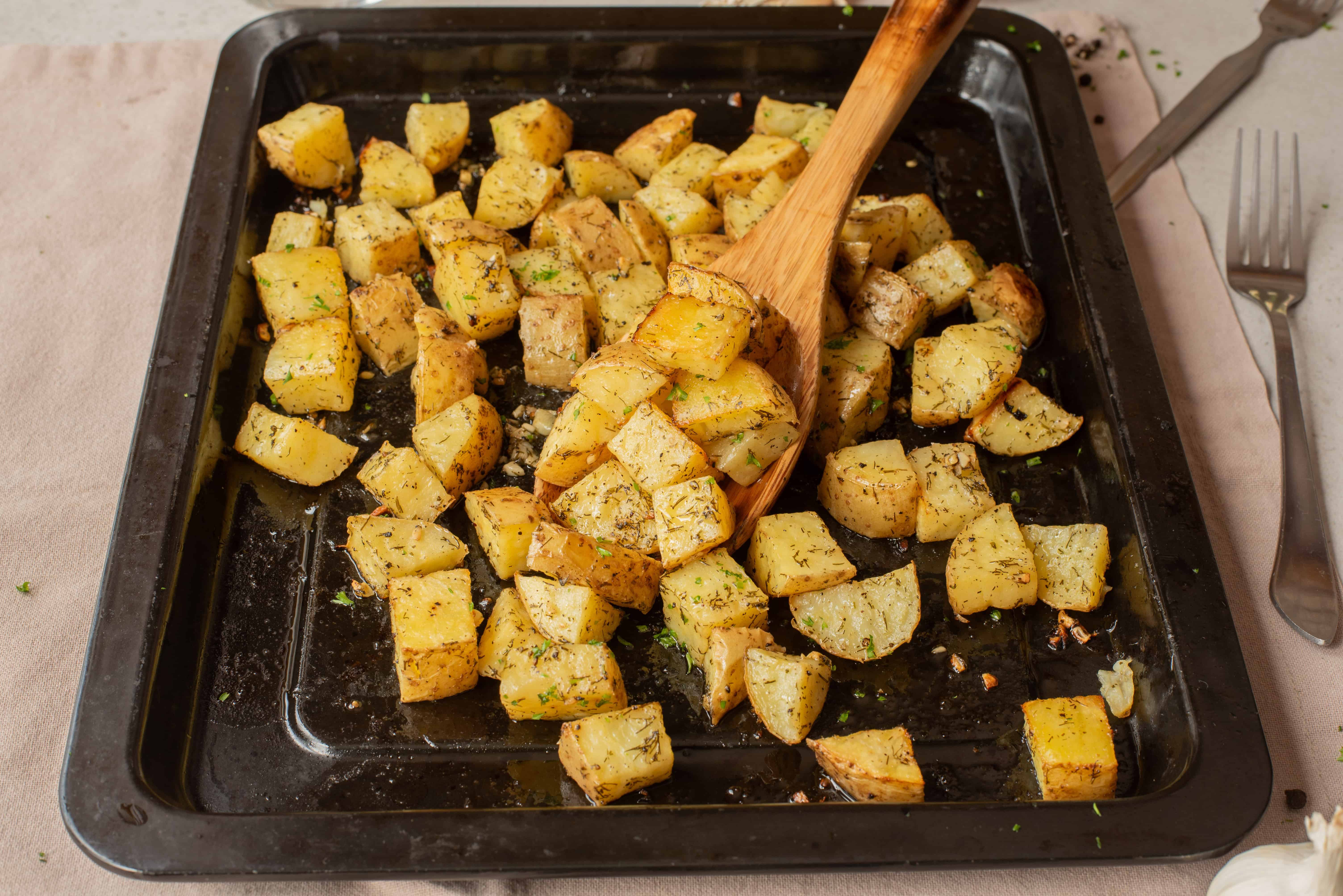 Roasted potato cubes with herbs on a baking tray, with a wooden spatula and cutlery on the side.