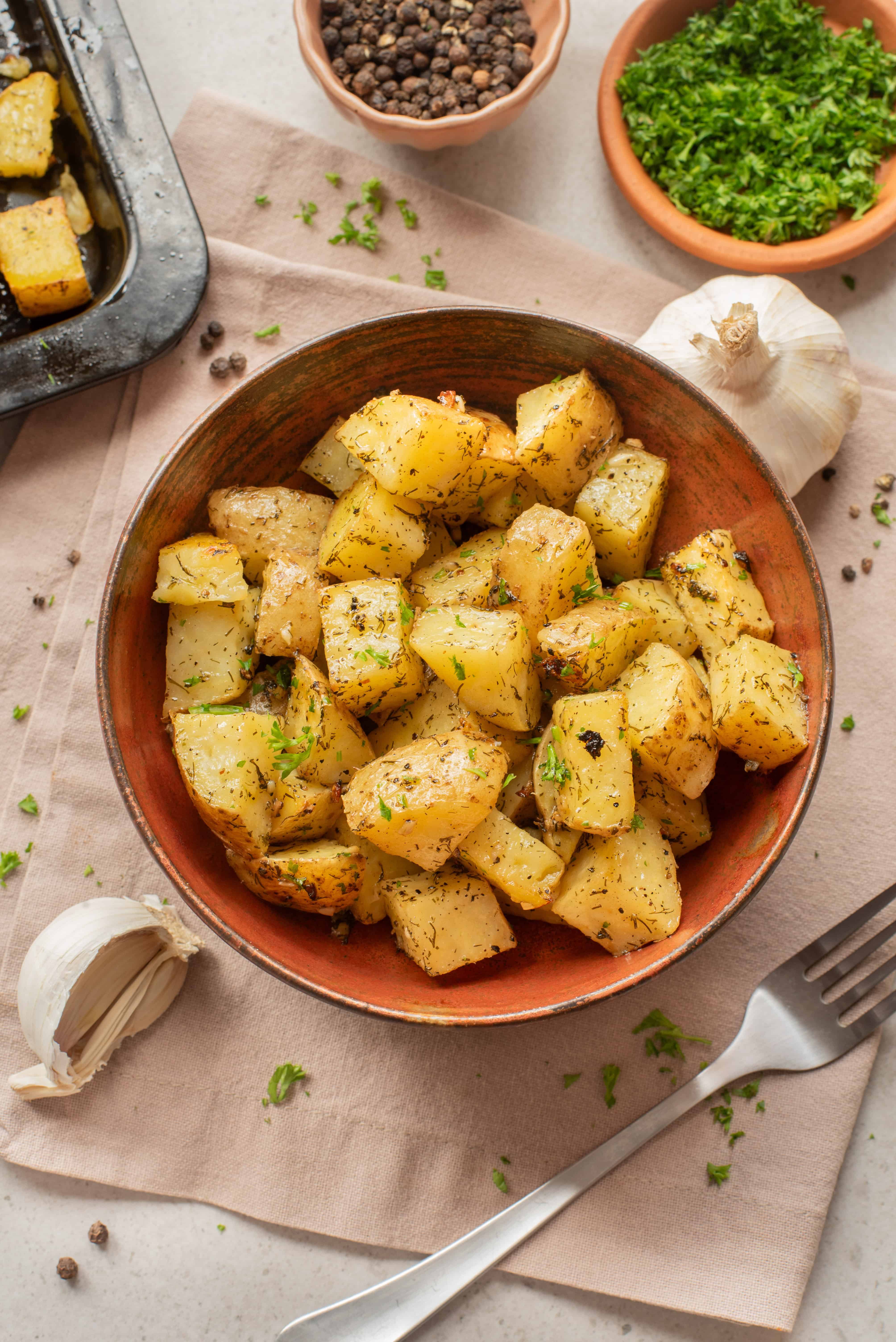 Bowl of roasted potatoes with herbs, surrounded by garlic, parsley, pepper, and a fork on a napkin.