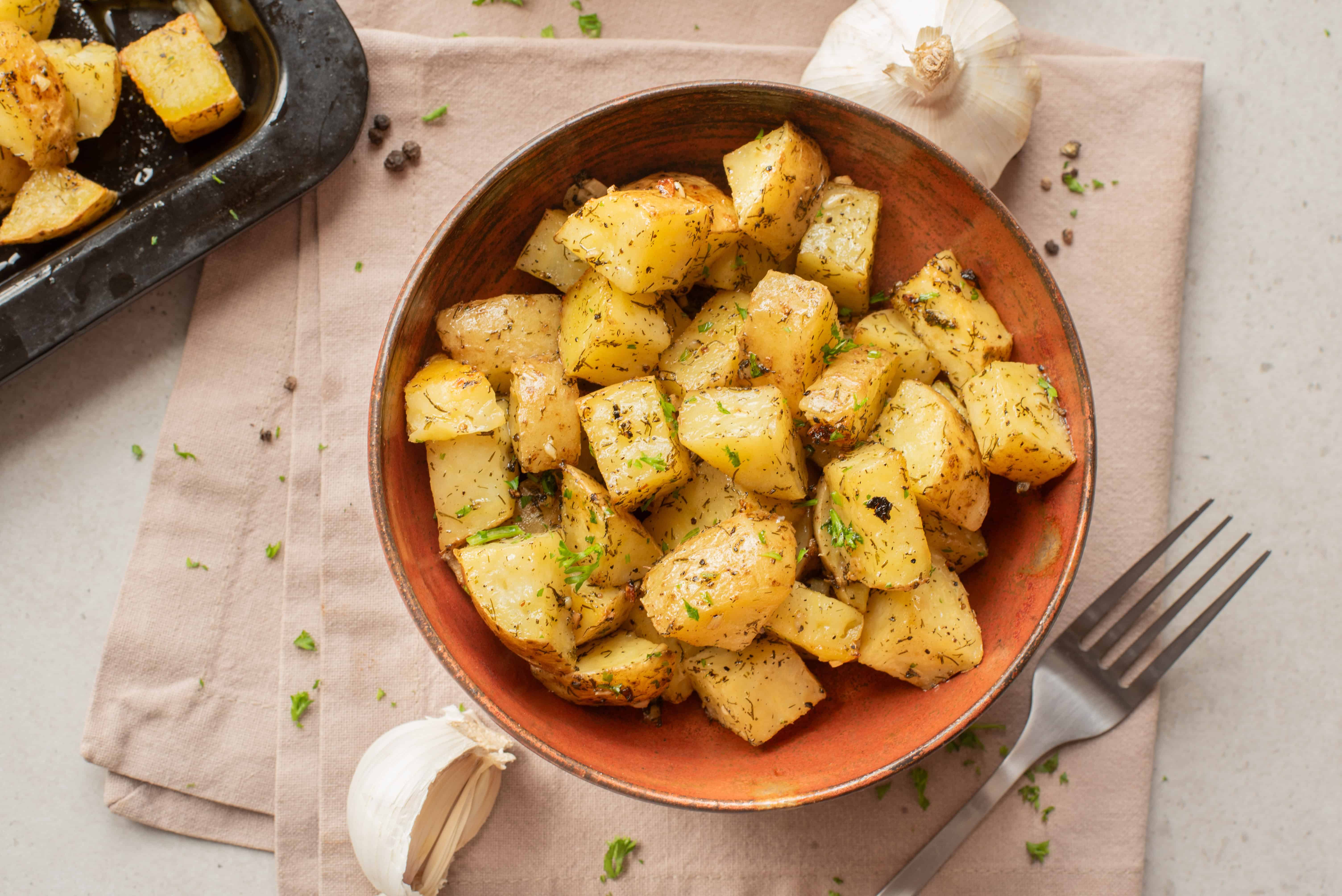 A bowl of roasted potatoes with herbs, next to a fork and garlic cloves on a beige napkin.
