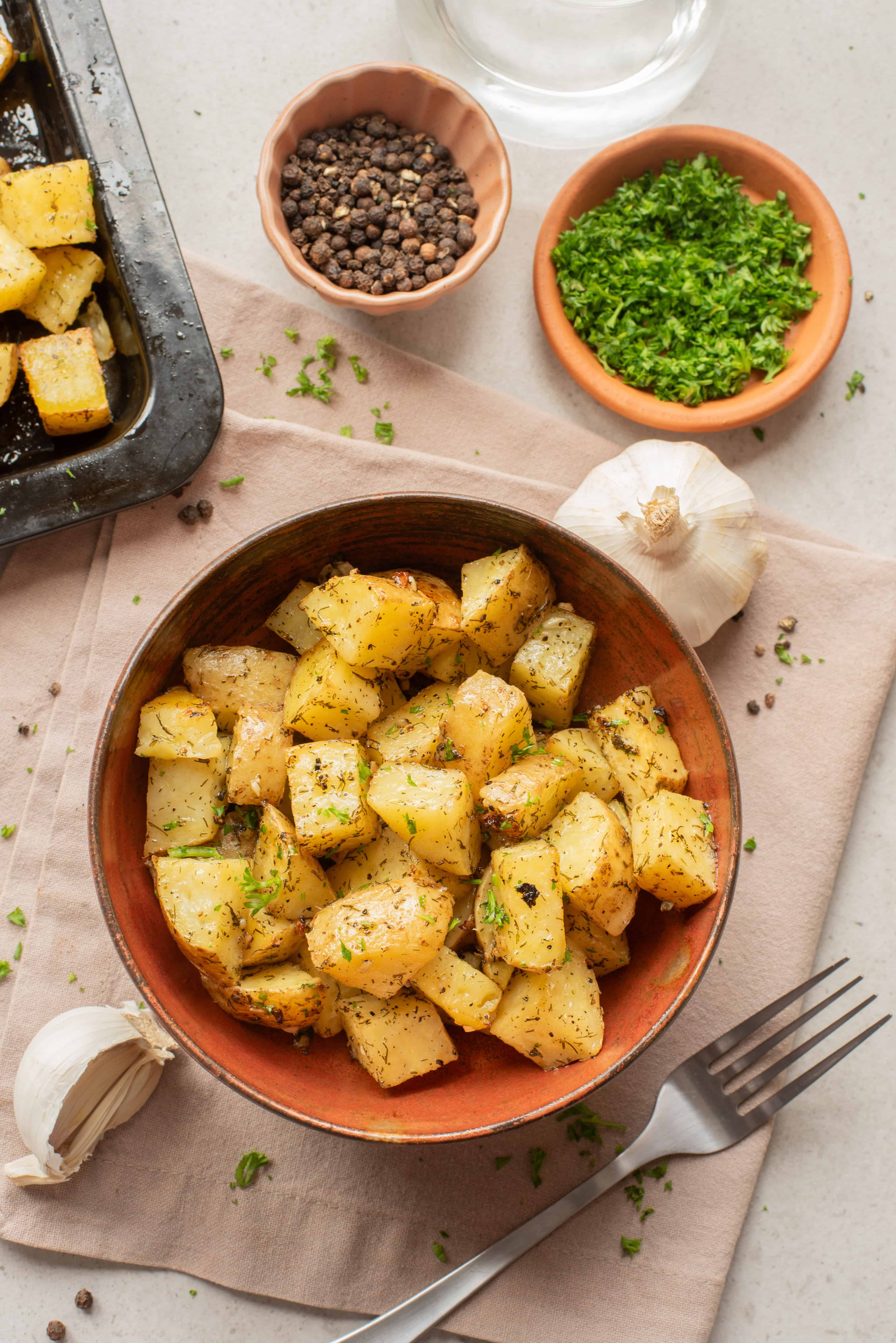 A bowl of roasted potatoes with herbs, surrounded by garlic, parsley, peppercorns, and a fork on a napkin.