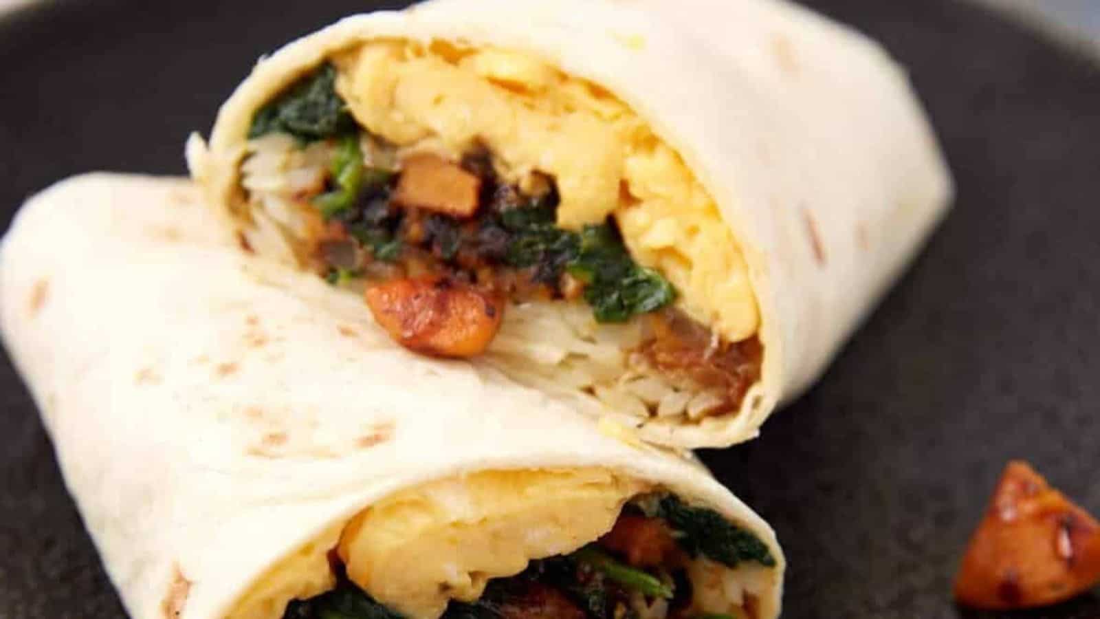 A breakfast burrito cut in half, filled with eggs, potatoes, spinach, and sausage on a dark plate.