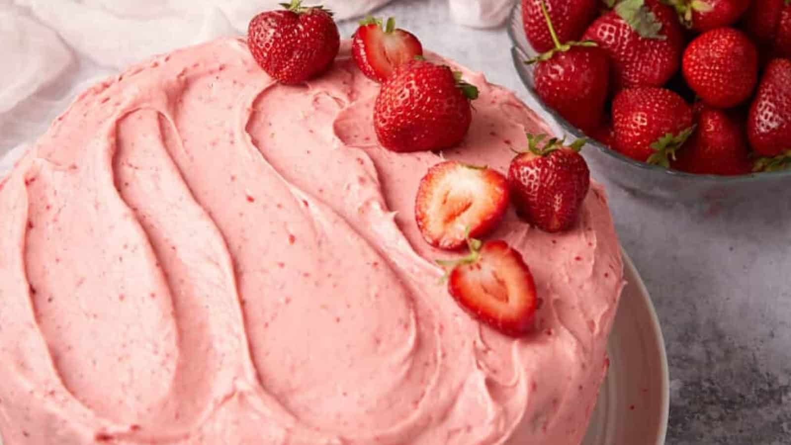 A frosted strawberry cake topped with fresh strawberries beside a bowl of whole strawberries.