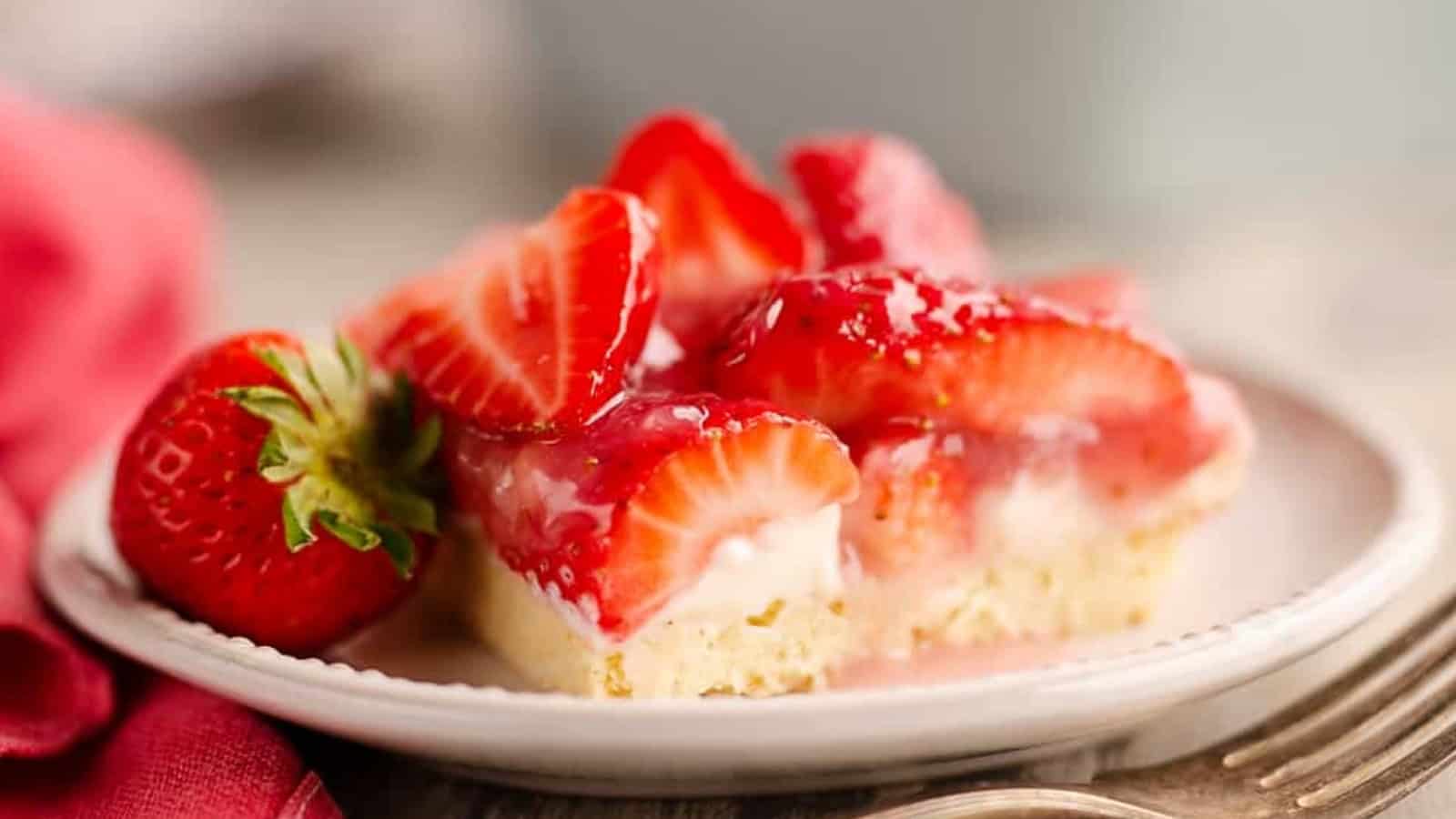 A slice of strawberry dessert bar with fresh strawberries and cream on a plate, next to a whole strawberry.