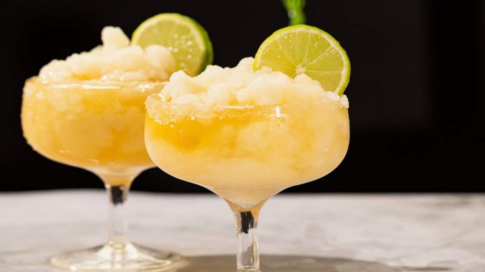 Two frozen margaritas in cocktail glasses with salt rims and lime slices, set against a dark background.