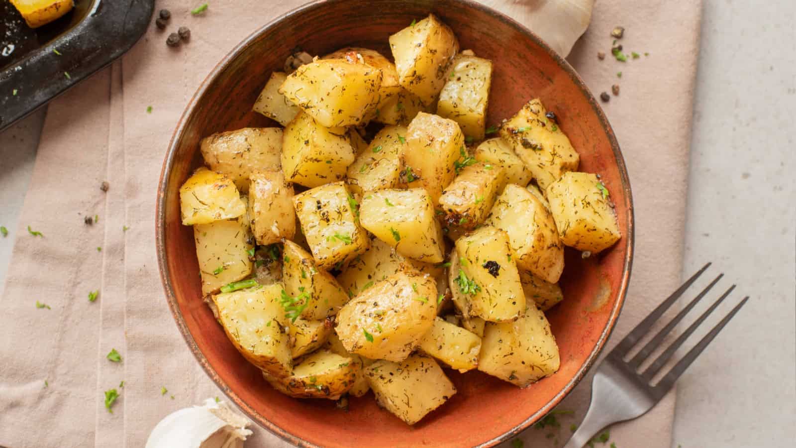 A bowl of seasoned roasted potatoes garnished with herbs, placed next to a fork and garlic cloves.