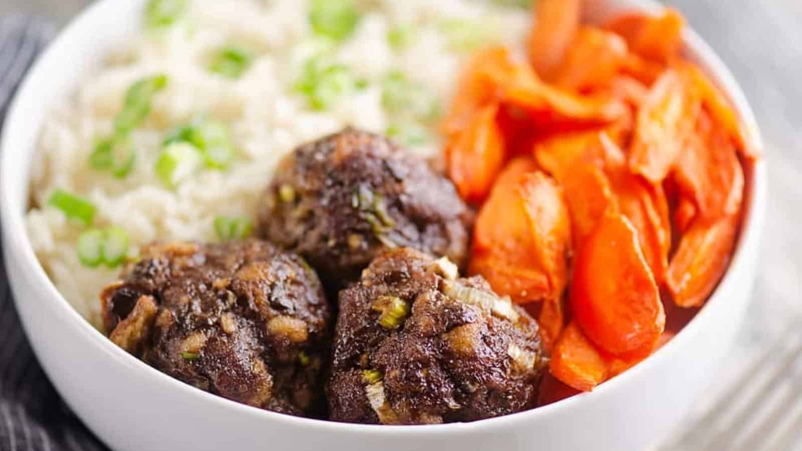 A bowl with rice topped with green onions, roasted carrots, and three meatballs.
