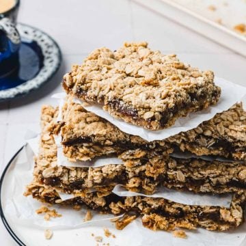 Stack of oat crumb bars with a jam filling on parchment paper, on a plate, with a cup in the background.