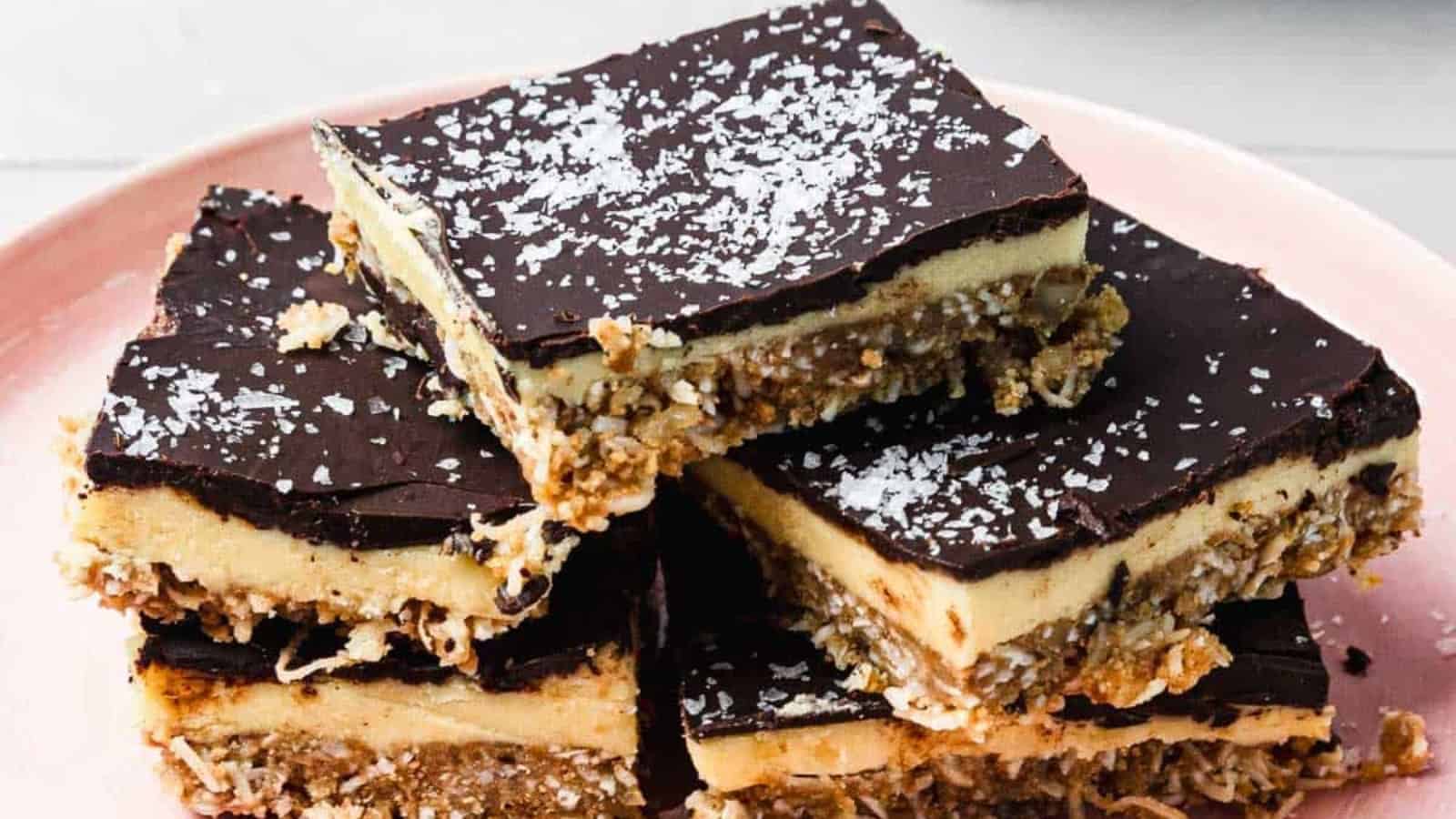 Stack of chocolate-topped dessert bars with creamy and nutty layers, sprinkled with shredded coconut.