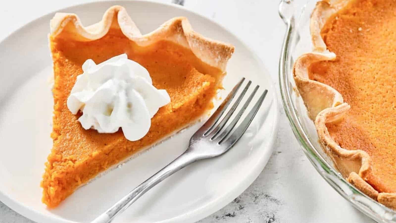 A slice of pumpkin pie with whipped cream on a plate, next to a fork and a pie dish with more pie.