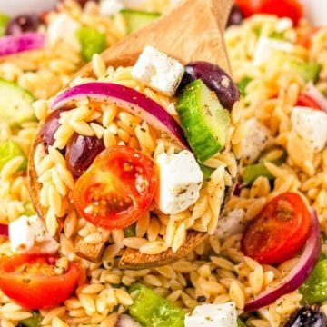 A wooden spoon scoops orzo pasta salad with tomatoes, cucumber, red onion, olives, and feta cheese.