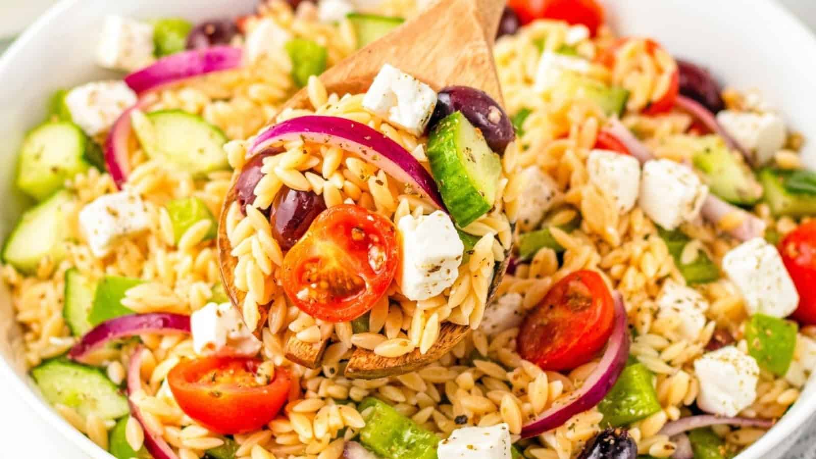A wooden spoon scoops orzo pasta salad with tomatoes, cucumber, red onion, olives, and feta cheese.