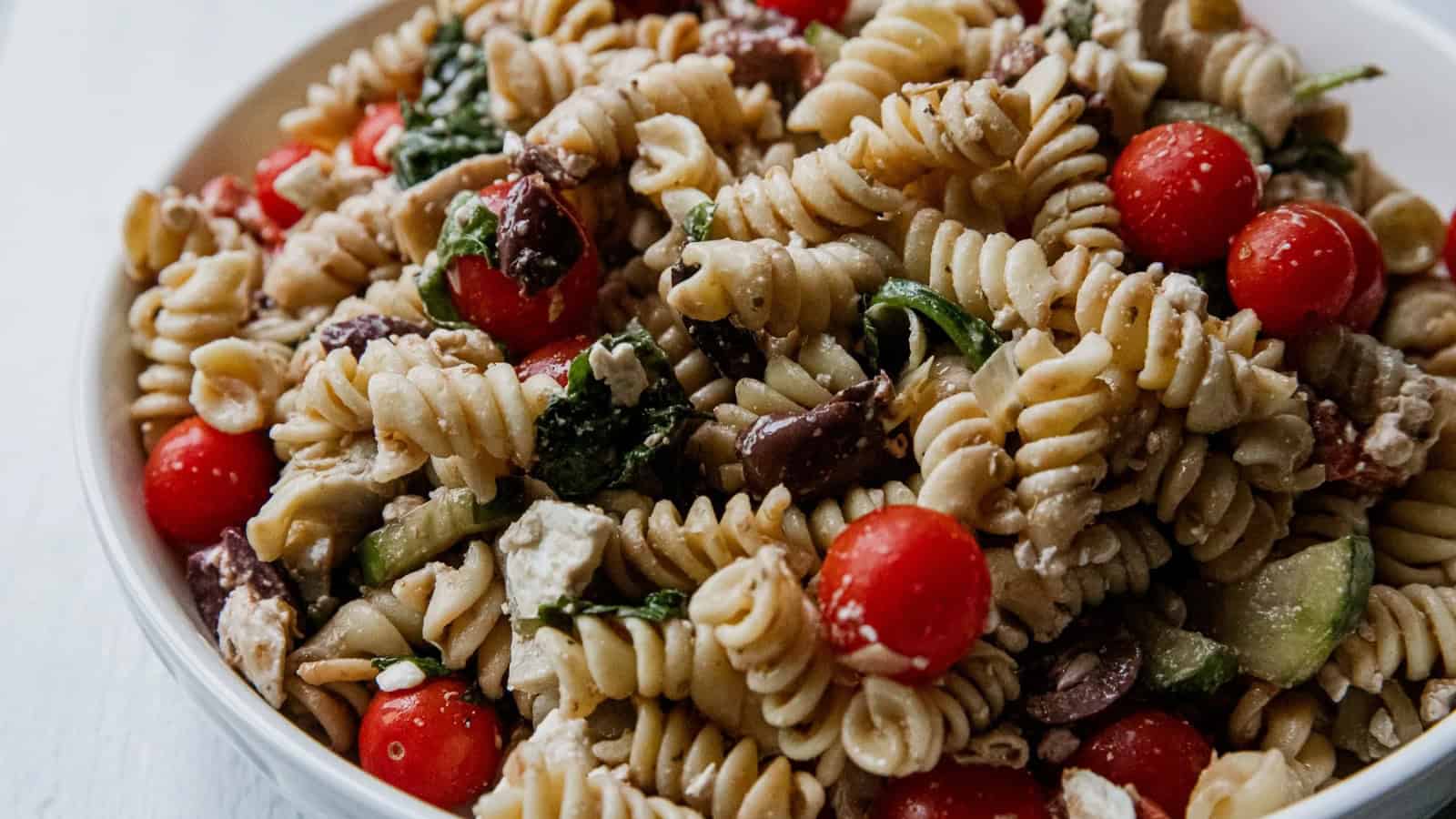 A bowl of rotini pasta salad with cherry tomatoes, spinach, feta cheese, and kalamata olives.