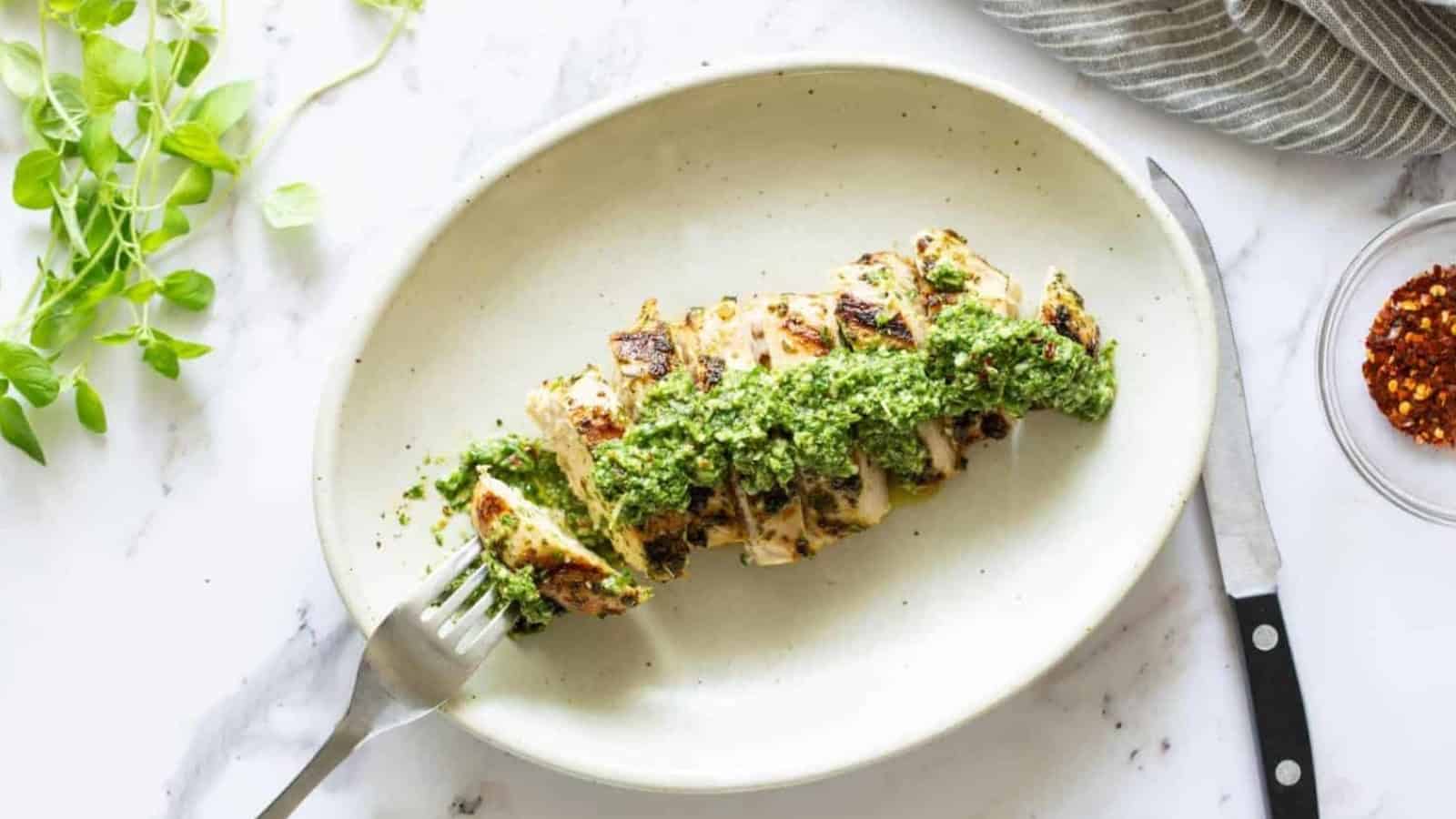 Sliced grilled chicken breast topped with green pesto sauce on an oval plate with a fork and knife.