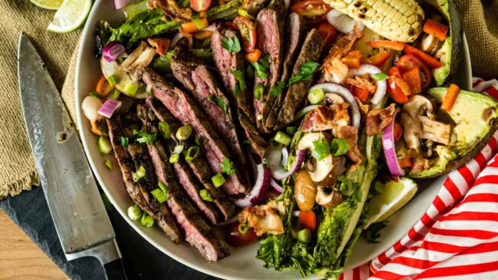 A bowl of grilled steak strips with vegetables, avocado, bacon, and corn beside a knife on a table.