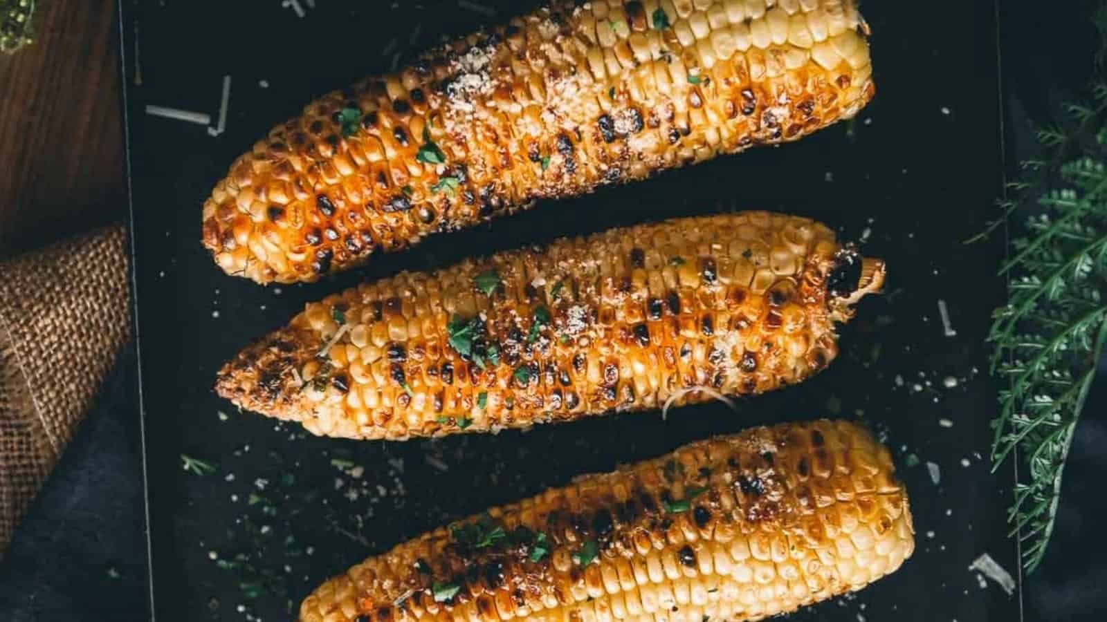 Three grilled corn cobs sprinkled with herbs and seasoning on a dark surface.
