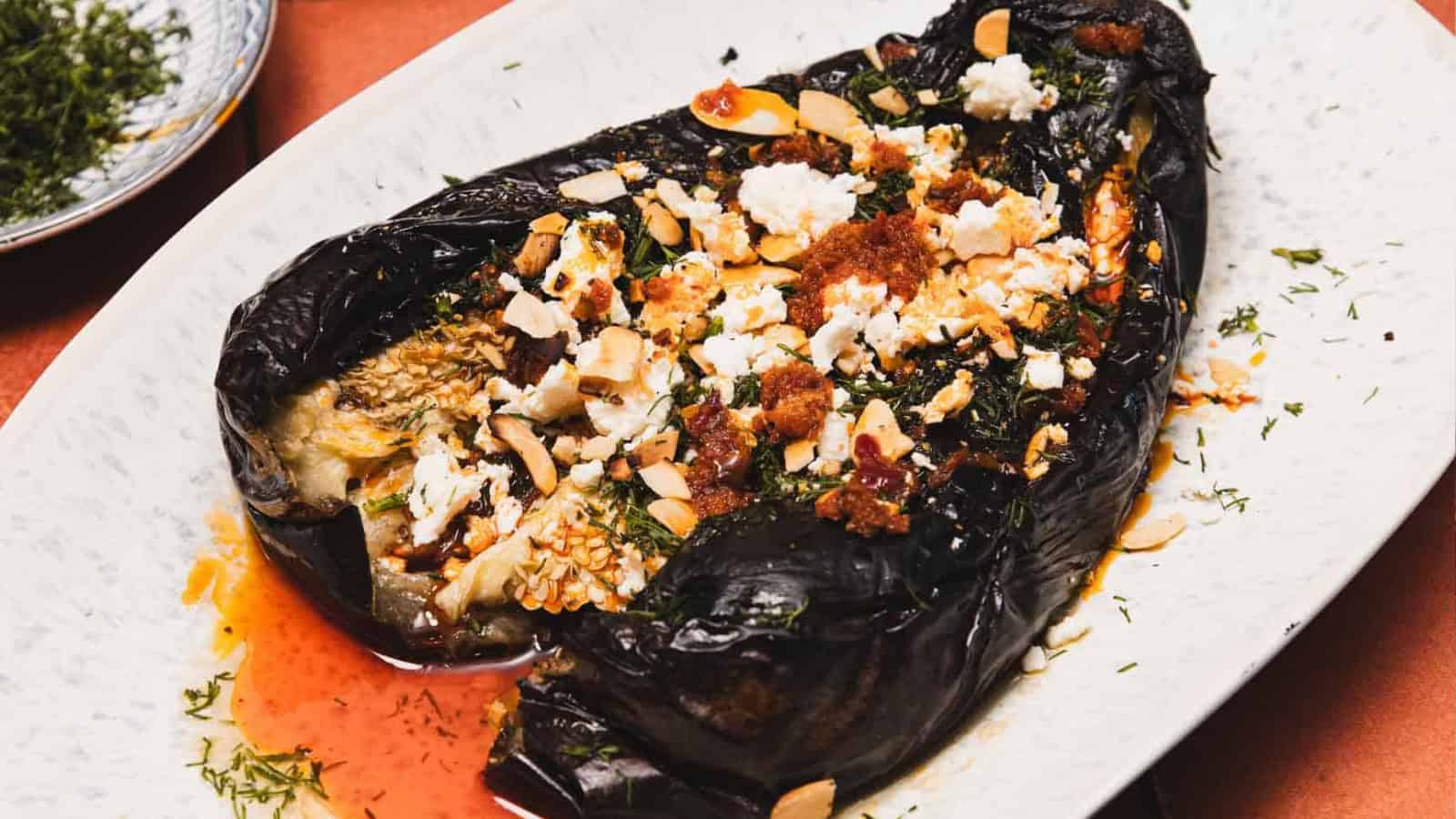 Charred stuffed eggplant topped with crumbled cheese, herbs, sliced almonds, and a drizzle of red oil on a plate.