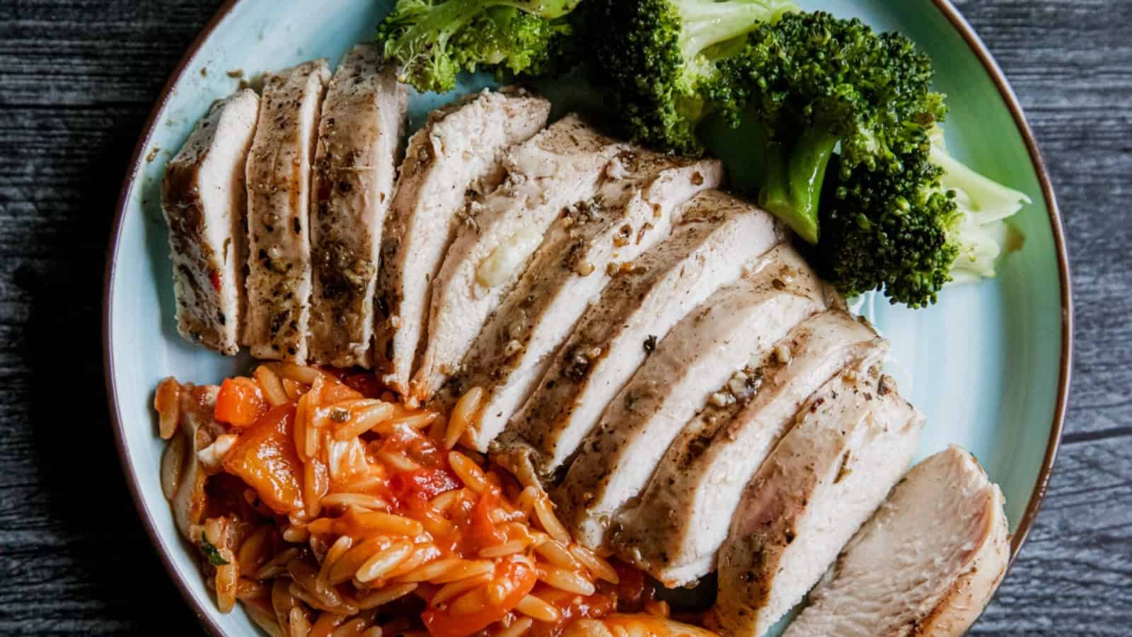Sliced grilled chicken breast with broccoli and tomato orzo on a blue plate.