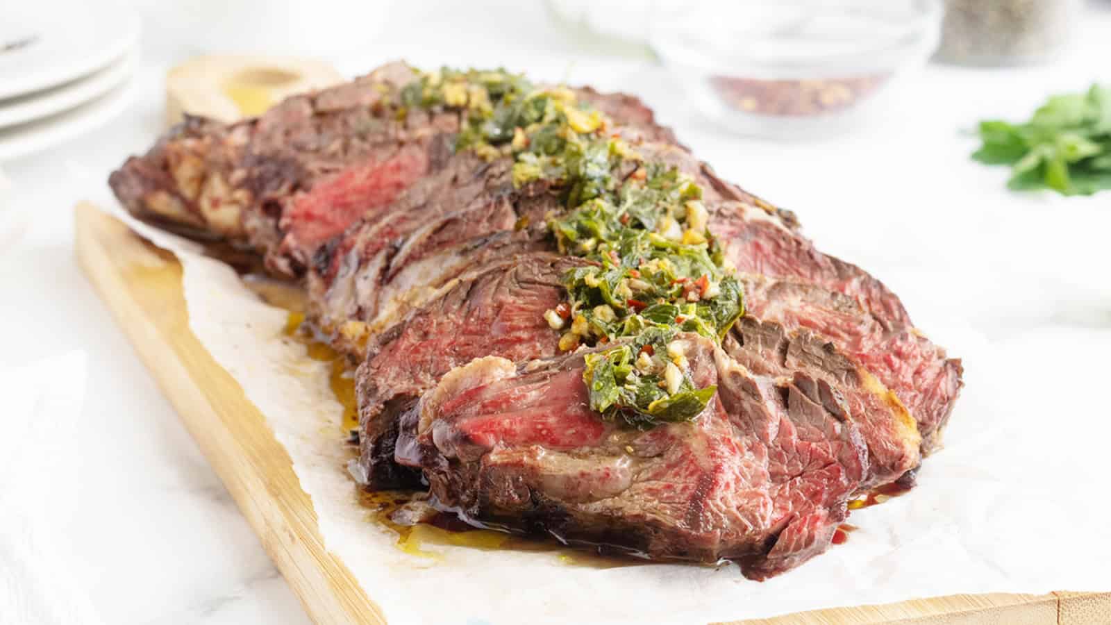 Sliced medium-rare steak topped with green chimichurri sauce on a wooden cutting board.