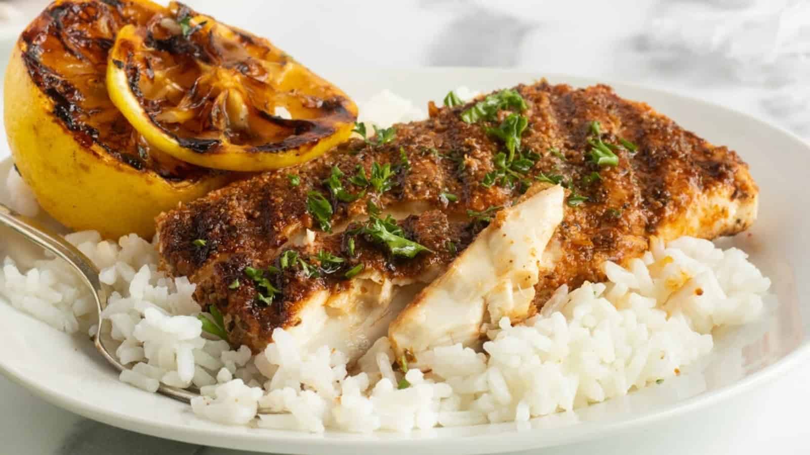 A plate of white rice topped with seasoned fish, fresh herbs, and a grilled lemon half.