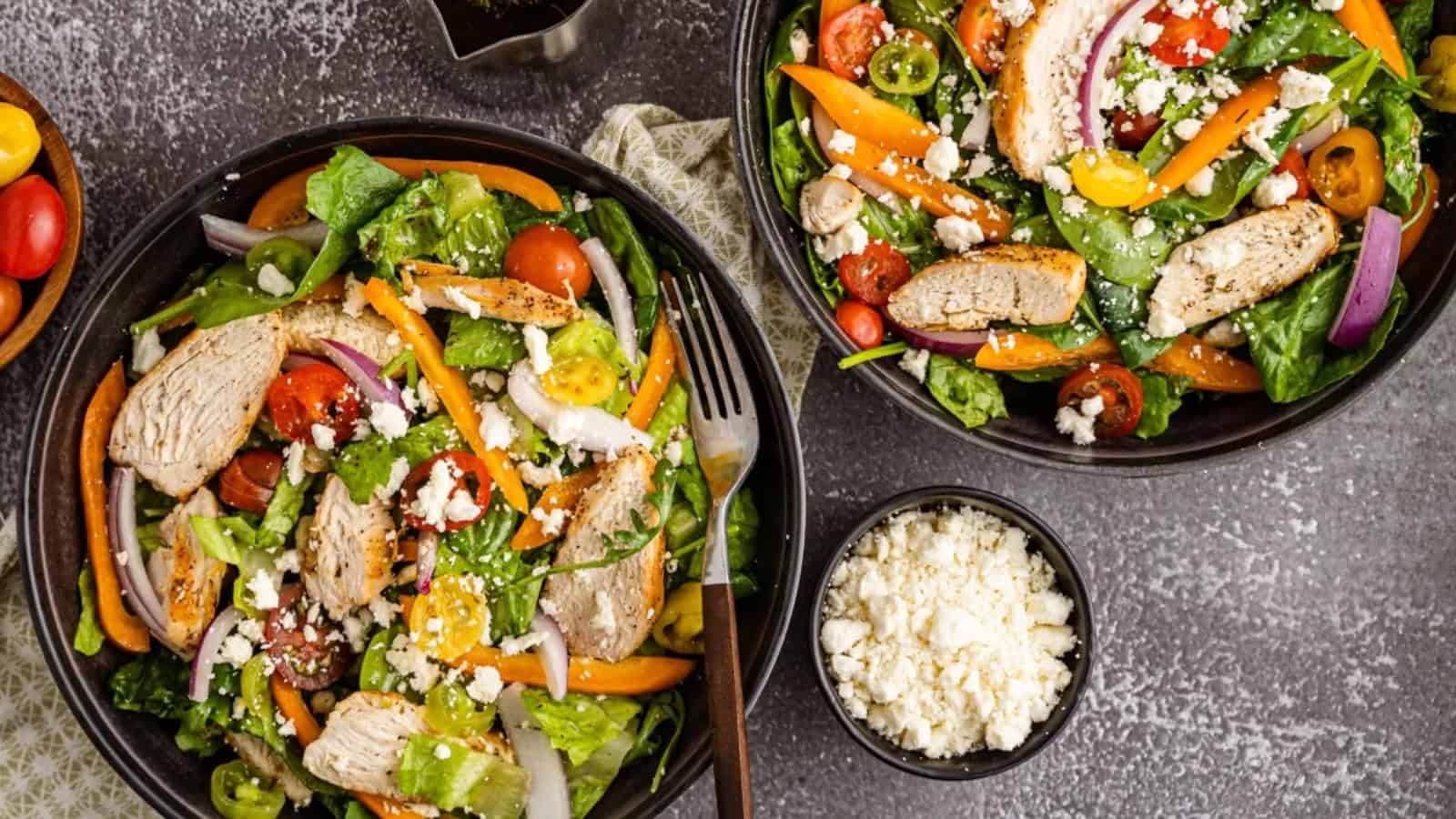 Two bowls of salad with grilled chicken, mixed greens, tomatoes, peppers, onions, and crumbled feta cheese.