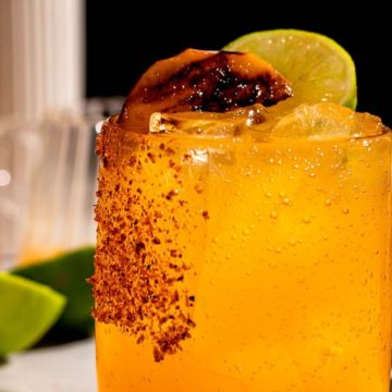 A cold, orange cocktail with ice, chili rim, grilled lime, and lime wedge, with bottles and limes in the background.
