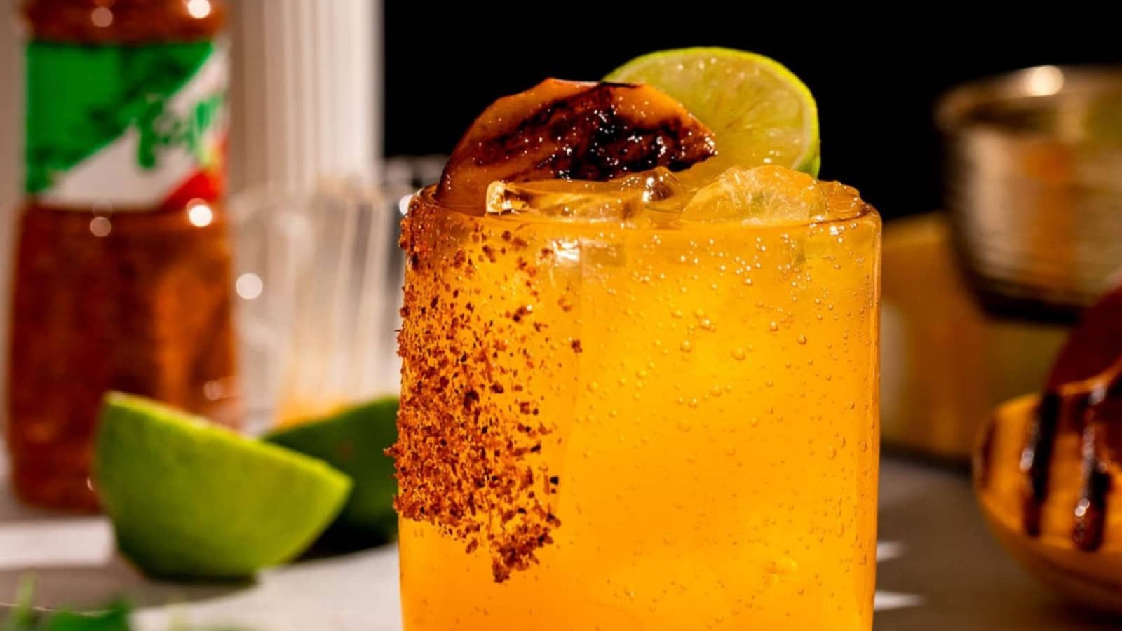 A cold, orange cocktail with ice, chili rim, grilled lime, and lime wedge, with bottles and limes in the background.