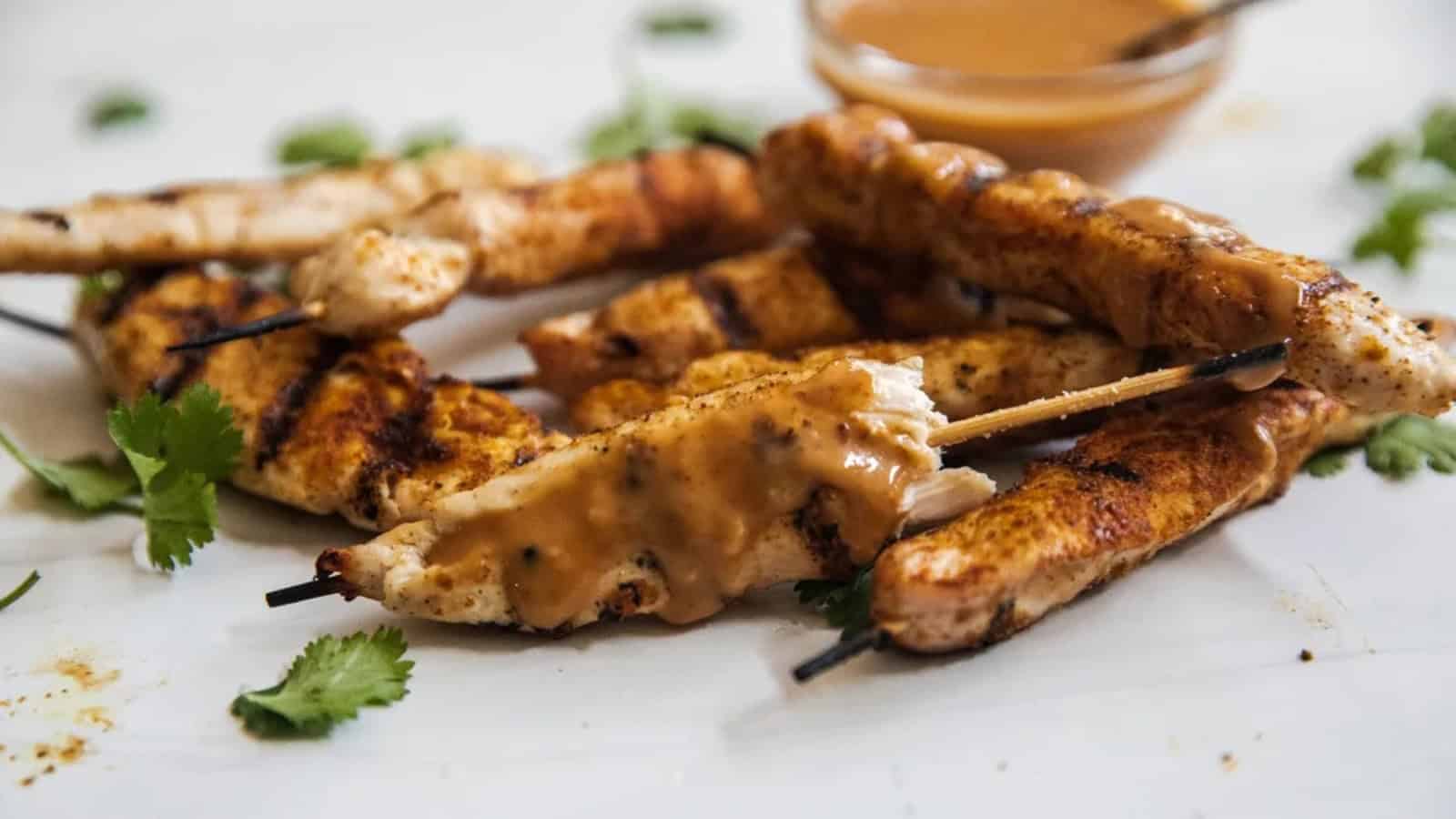 Grilled chicken skewers topped with peanut sauce, garnished with fresh cilantro on a white surface.